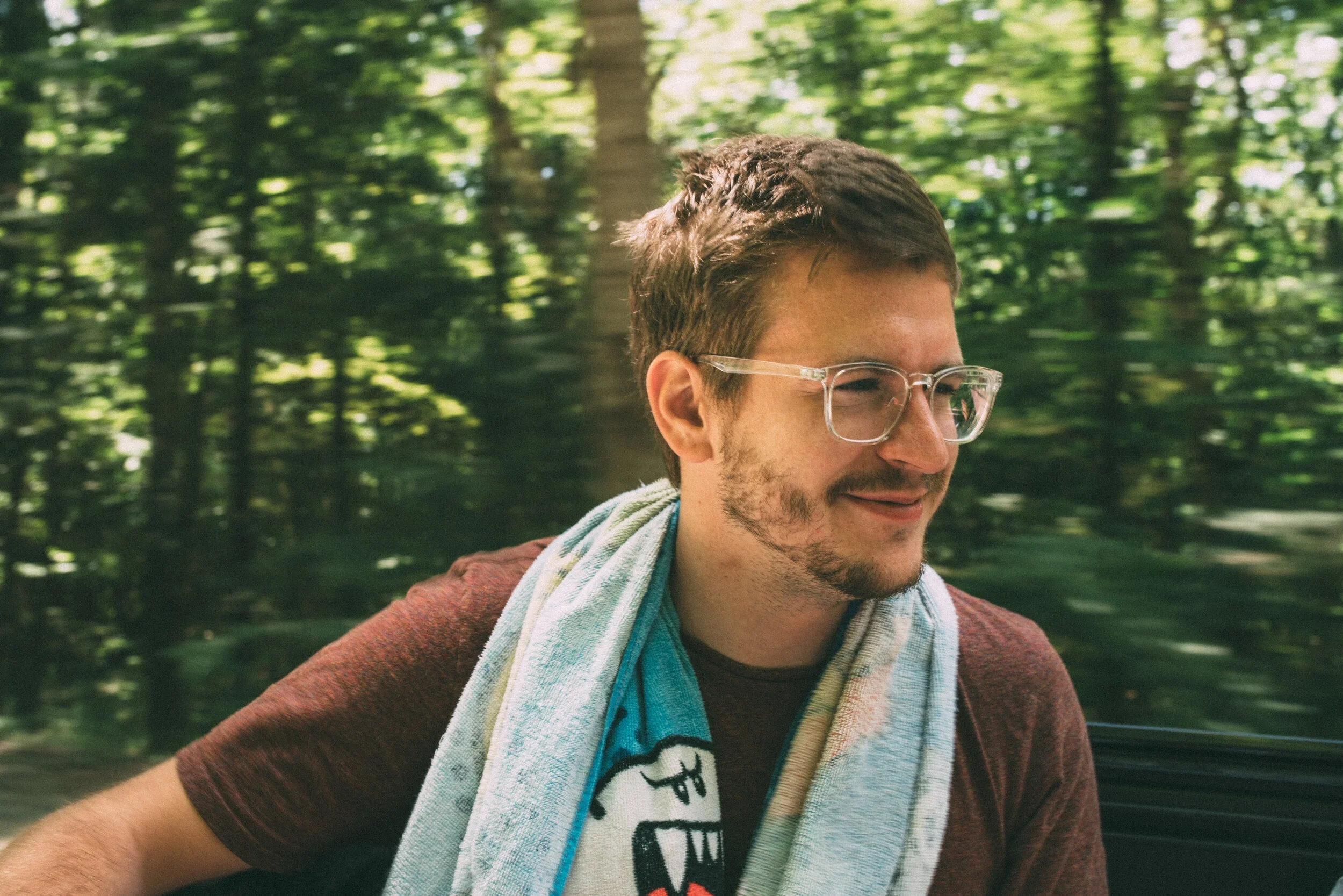 A man with glasses and a beard smiling, sitting outdoors with trees in the background, wearing a maroon shirt and a towel around his neck.