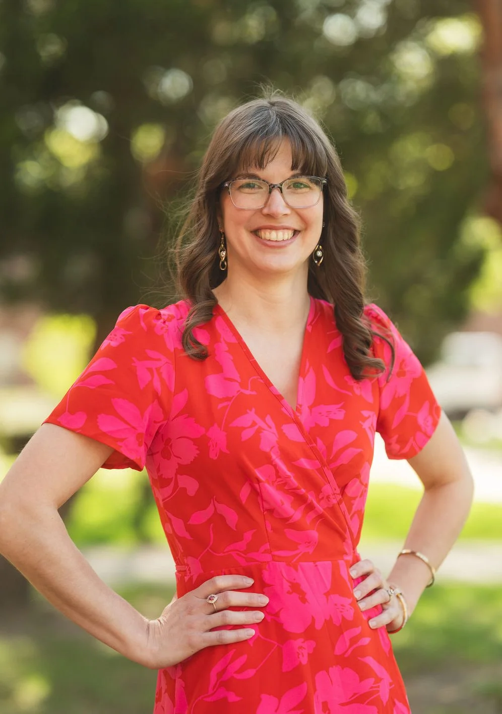 A woman with glasses and long brown hair smiling outdoors, wearing a pink floral dress and gold jewelry.