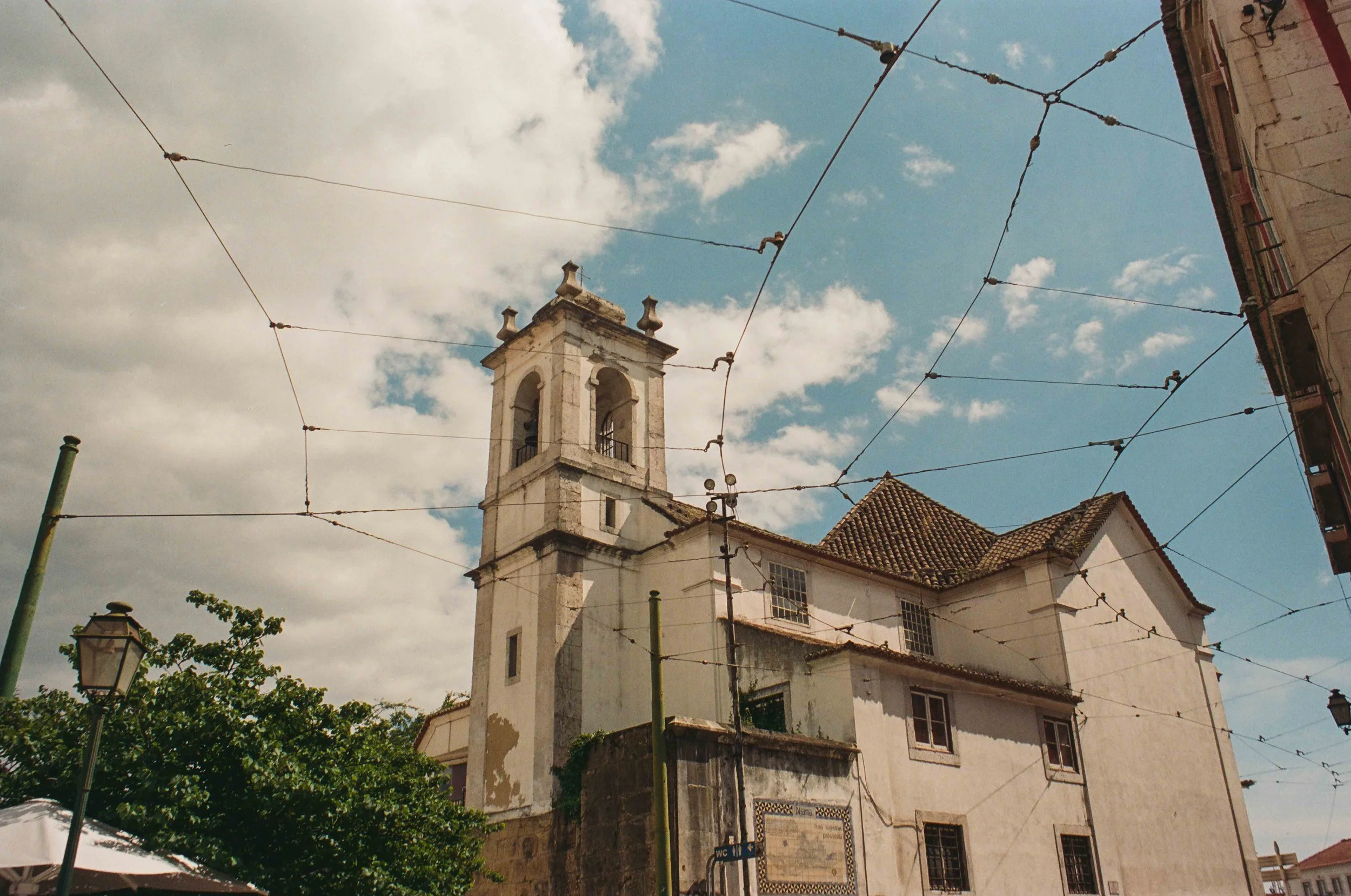 A historic church with a bell tower and tiled roof, under a partly cloudy sky, with overhead tram wires and a streetlamp in the foreground.