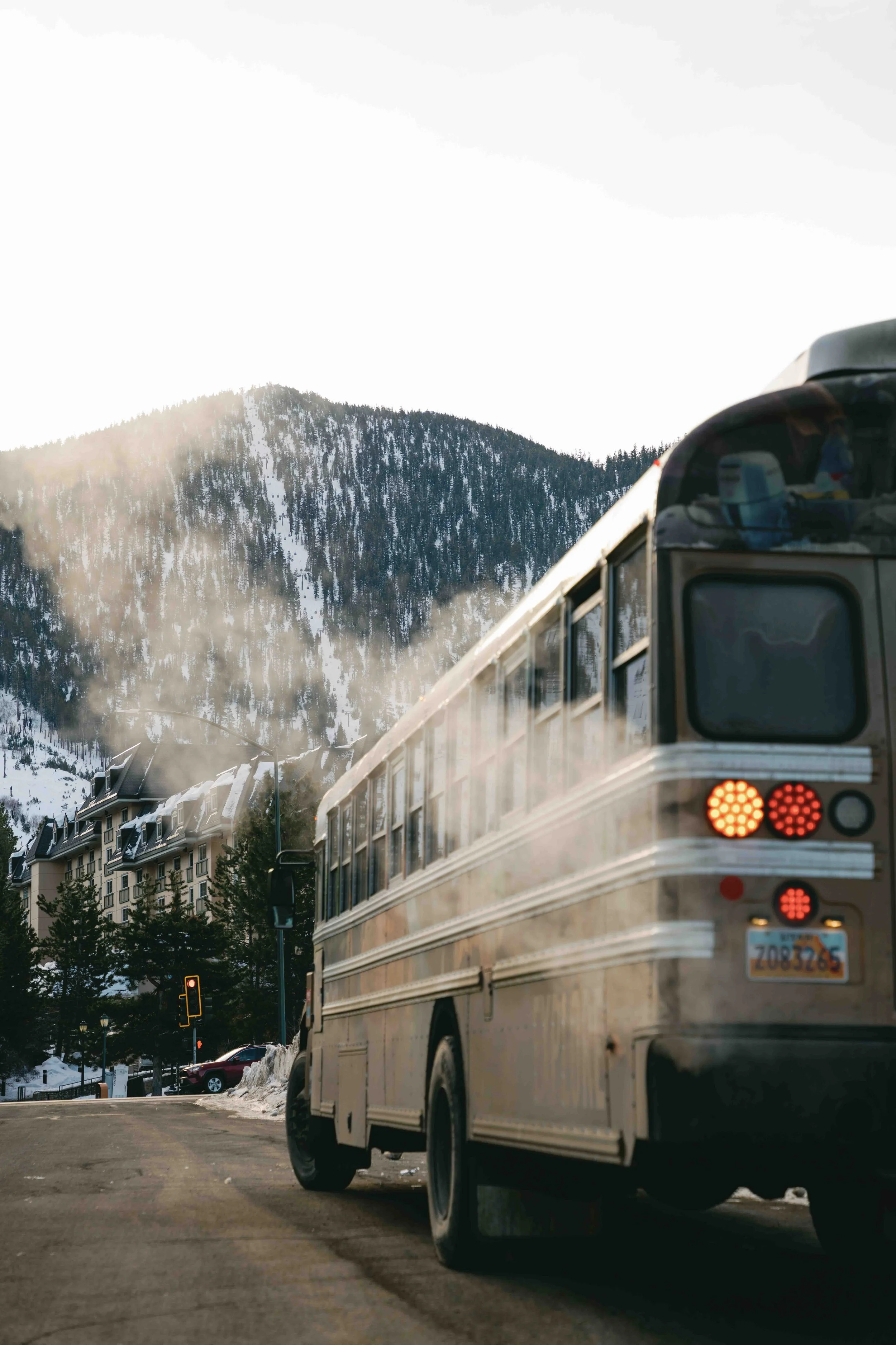 A city street with buildings, trees, and traffic lights, with a large mountain in the background. A bus is stopped, with steam or exhaust visible around it.