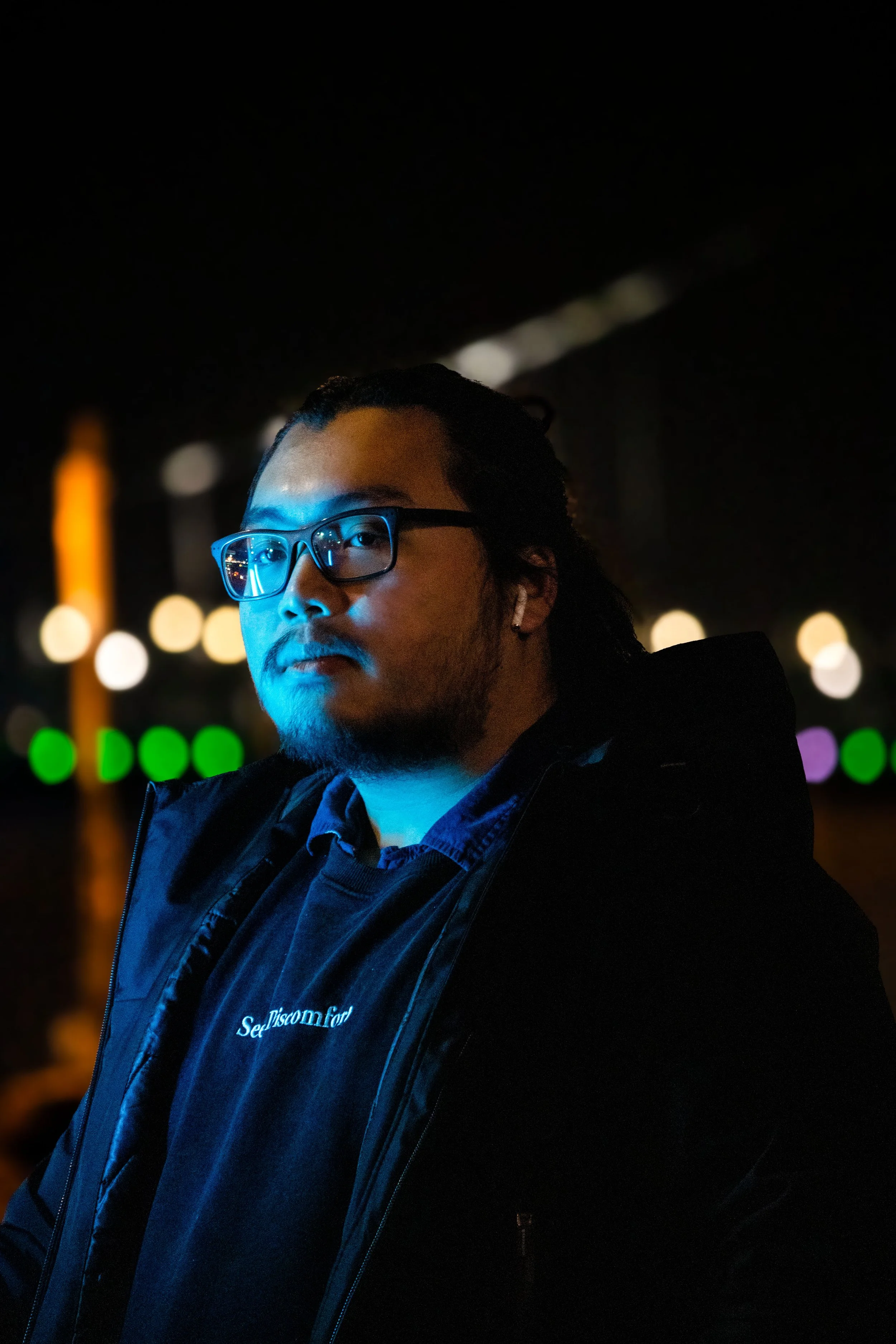 A man with glasses and facial hair stands outdoors at night, illuminated by blue and orange lights, with blurred colorful city lights in the background.