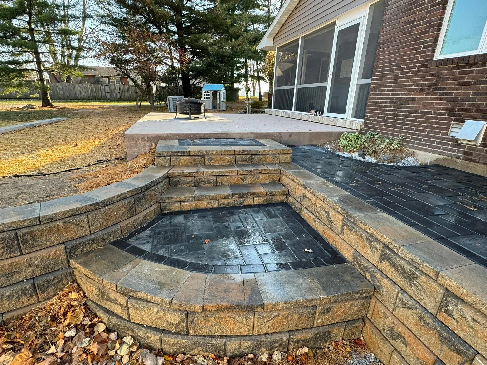 Backyard with a brick and stone staircase leading to a higher patio area. The patio has outdoor furniture and a children's playhouse. The yard is partially landscaped with trees and a fence in the background.