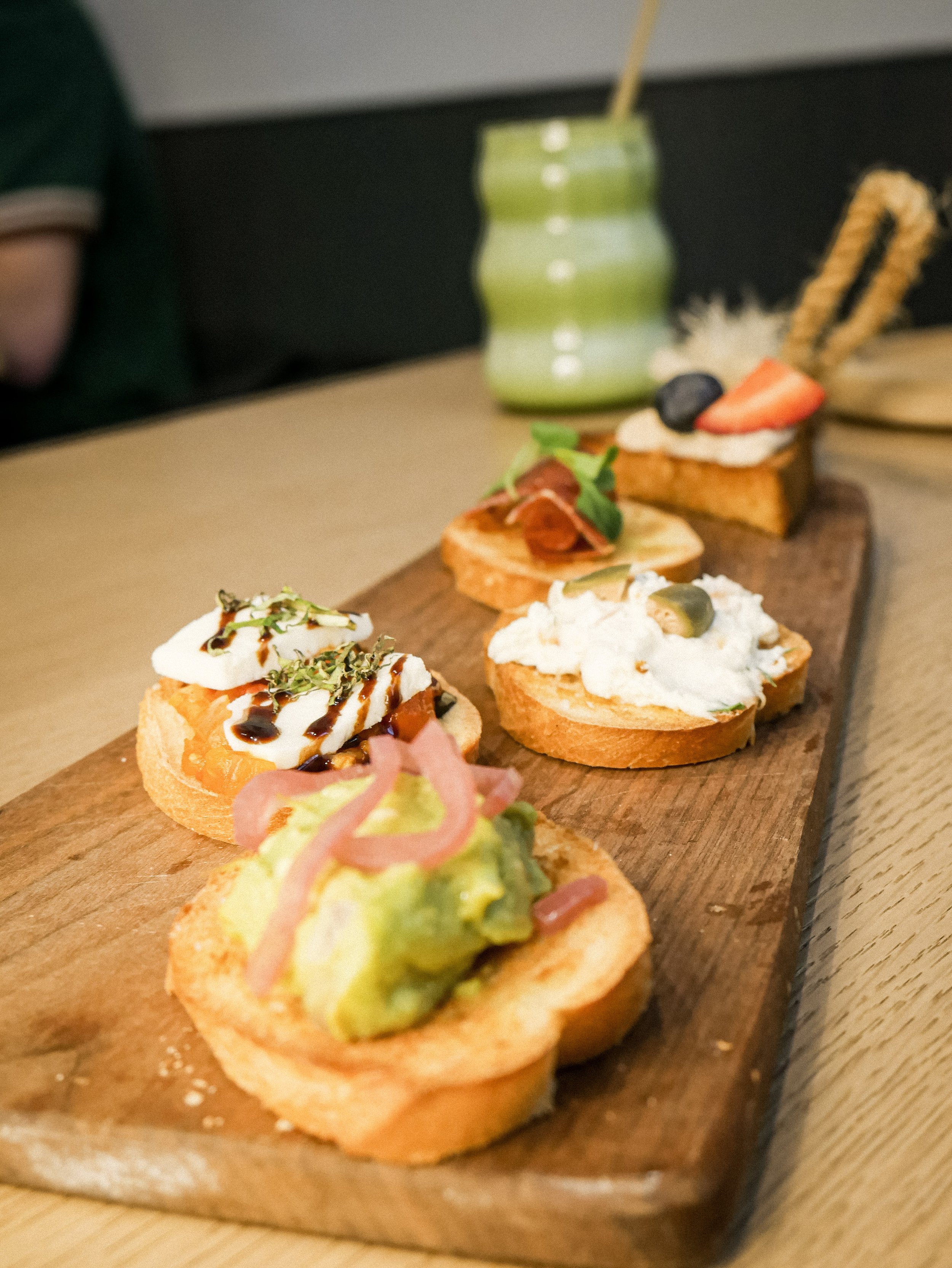 A wooden serving board with six different types of tapas or canapés, each topped with various ingredients, arranged in a line. In the background, there is a green striped jar with a straw and a person partially visible.