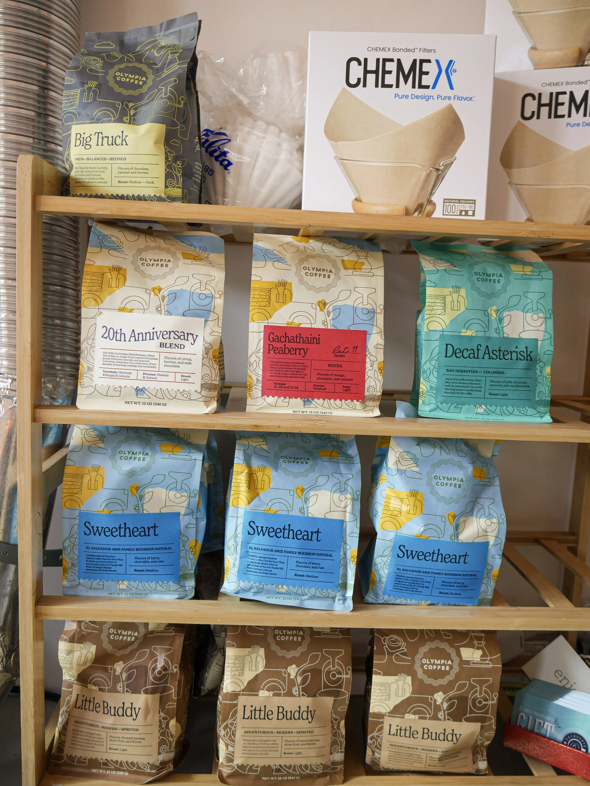 A wooden shelf holding five different bags of Olympia Coffee with various colorful labels, ranging from top to bottom: Big Truck, 20th Anniversary Blend, Gachathaini Peaberry, Decaf Asterisk, and three bags of Sweetheart and three bags of Little Budd