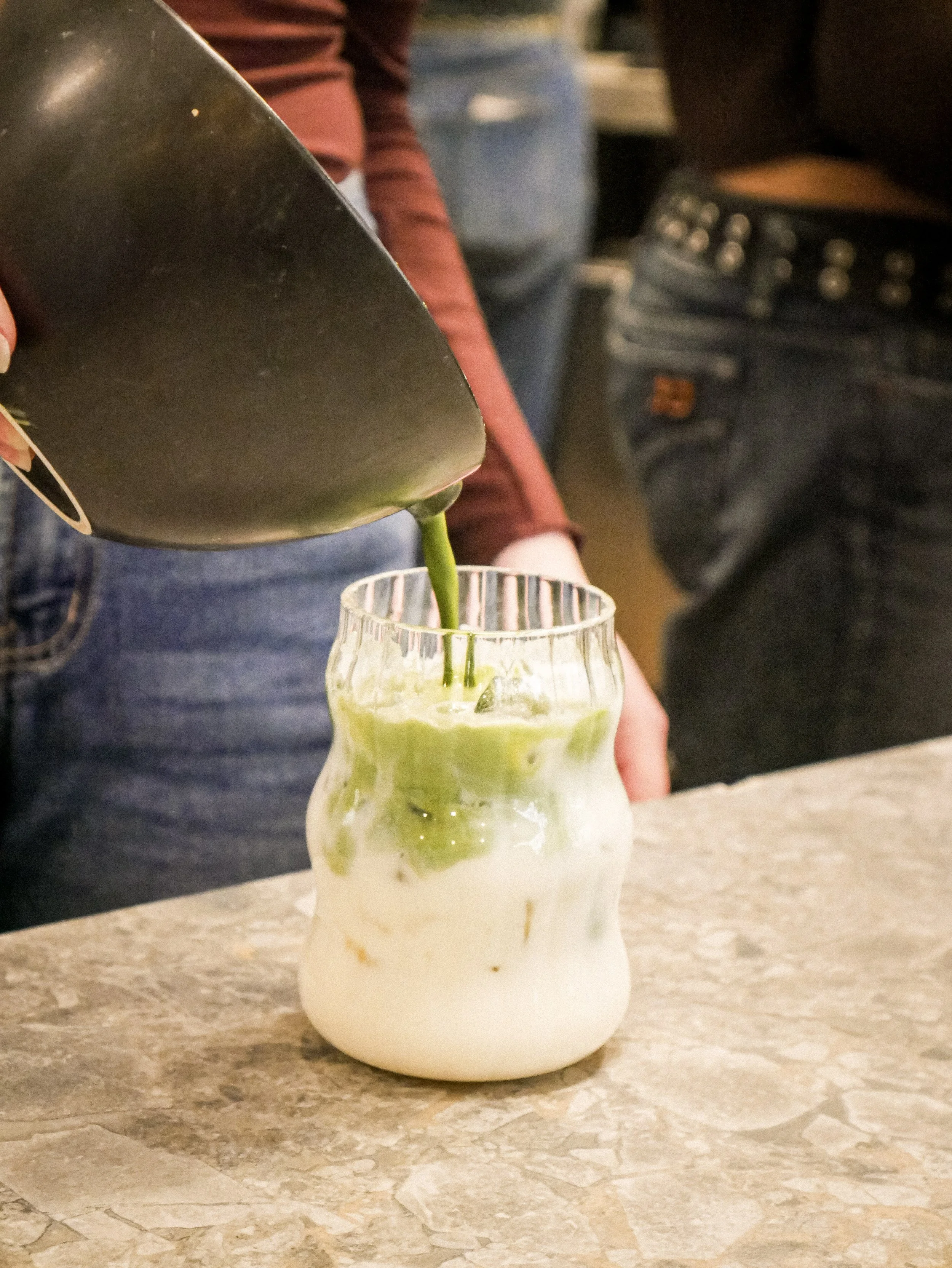 Person pouring green matcha latte into a clear glass with a wavy design on a marble countertop.