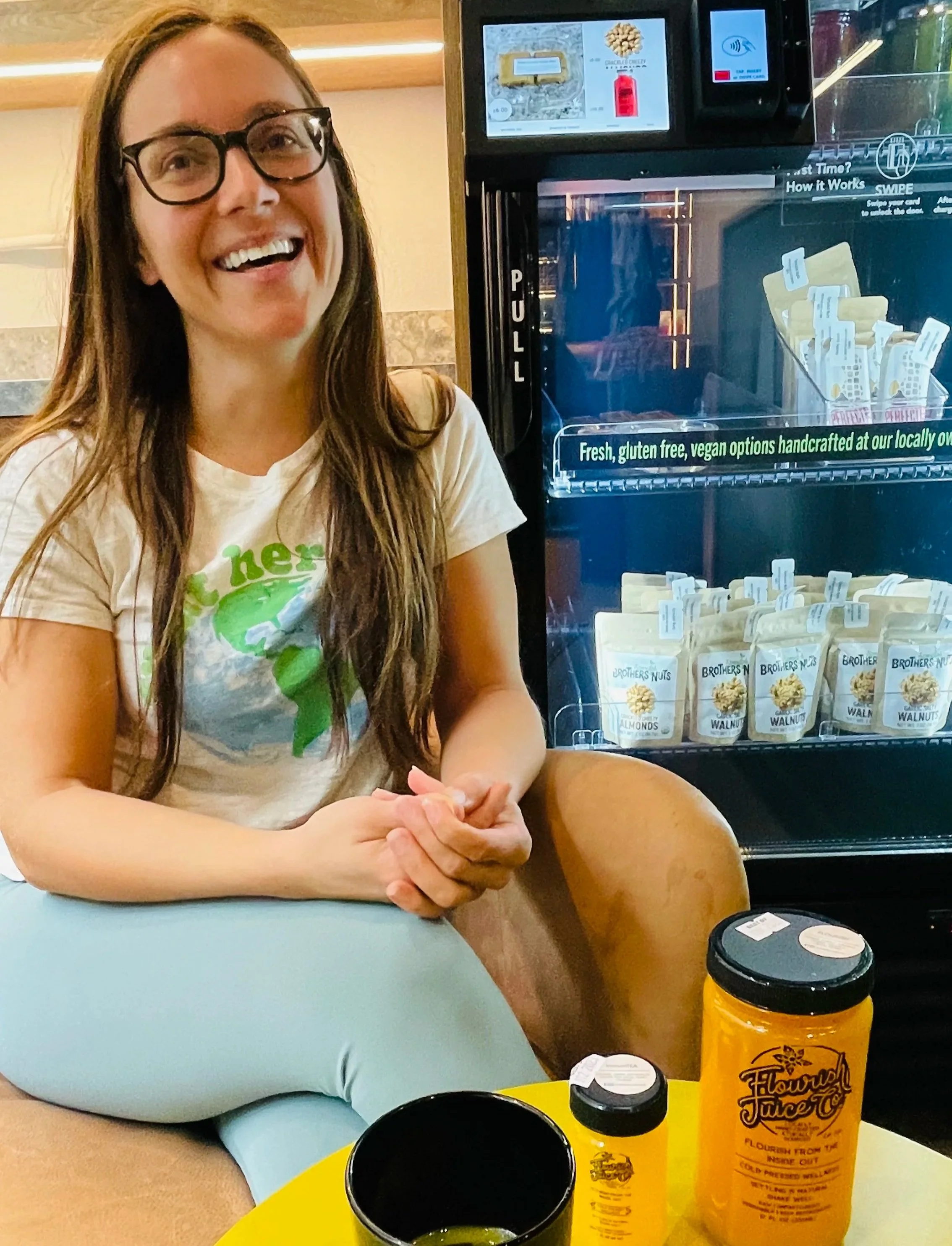 A woman with long brown hair and glasses smiling, sitting at a table with a yellow juice bottle and a black cup in front of her. Behind her is a refrigerator with packaged nuts. Kaitlin Soto of Flourish Juice Company and the Byte automated store.