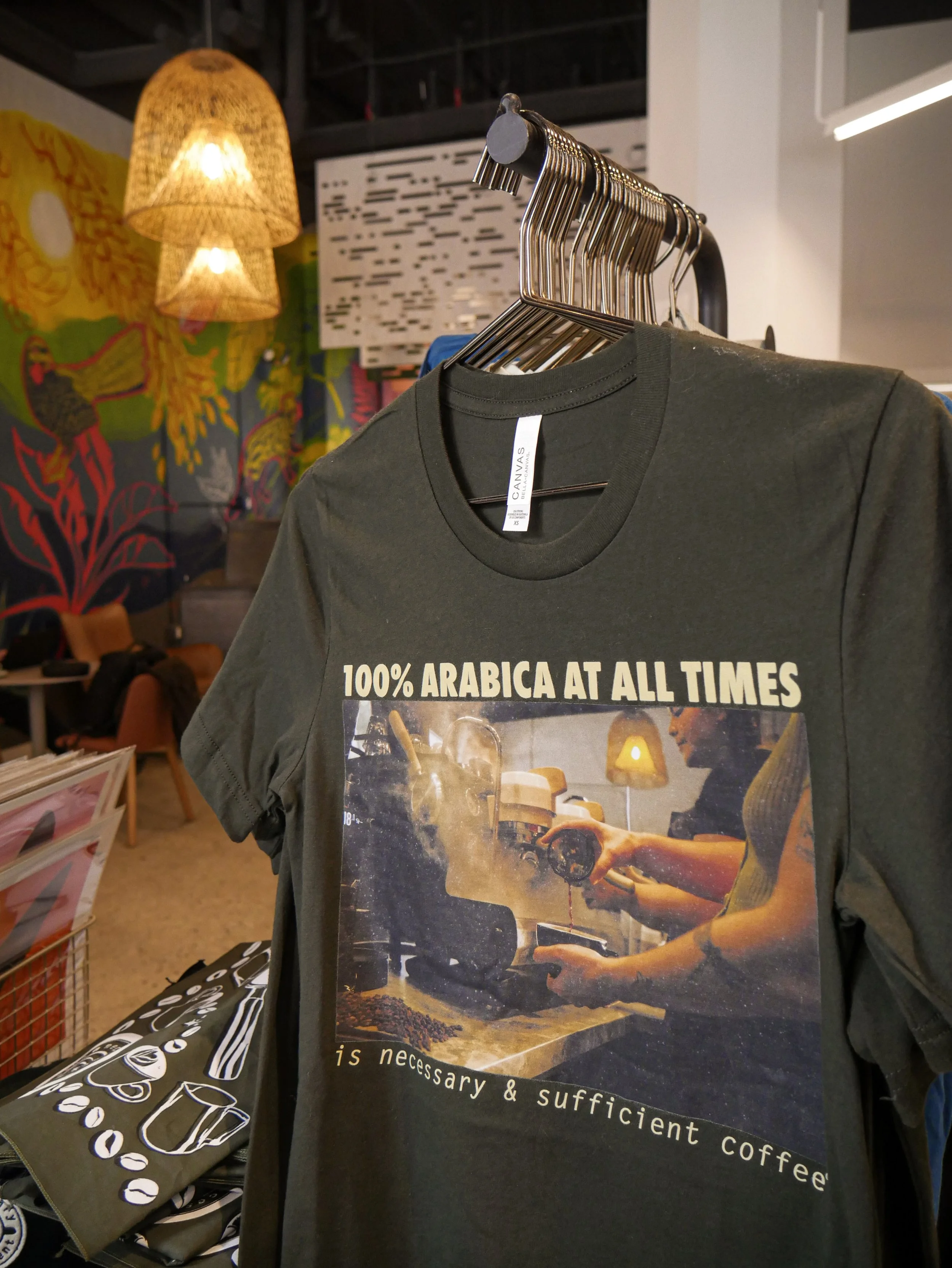 A dark green T-shirt hanging on a clothing rack with text that reads "100% Arabica at all times is necessary & sufficient coffee." The T-shirt features an image of baristas preparing coffee, with a warm, cozy cafe background.