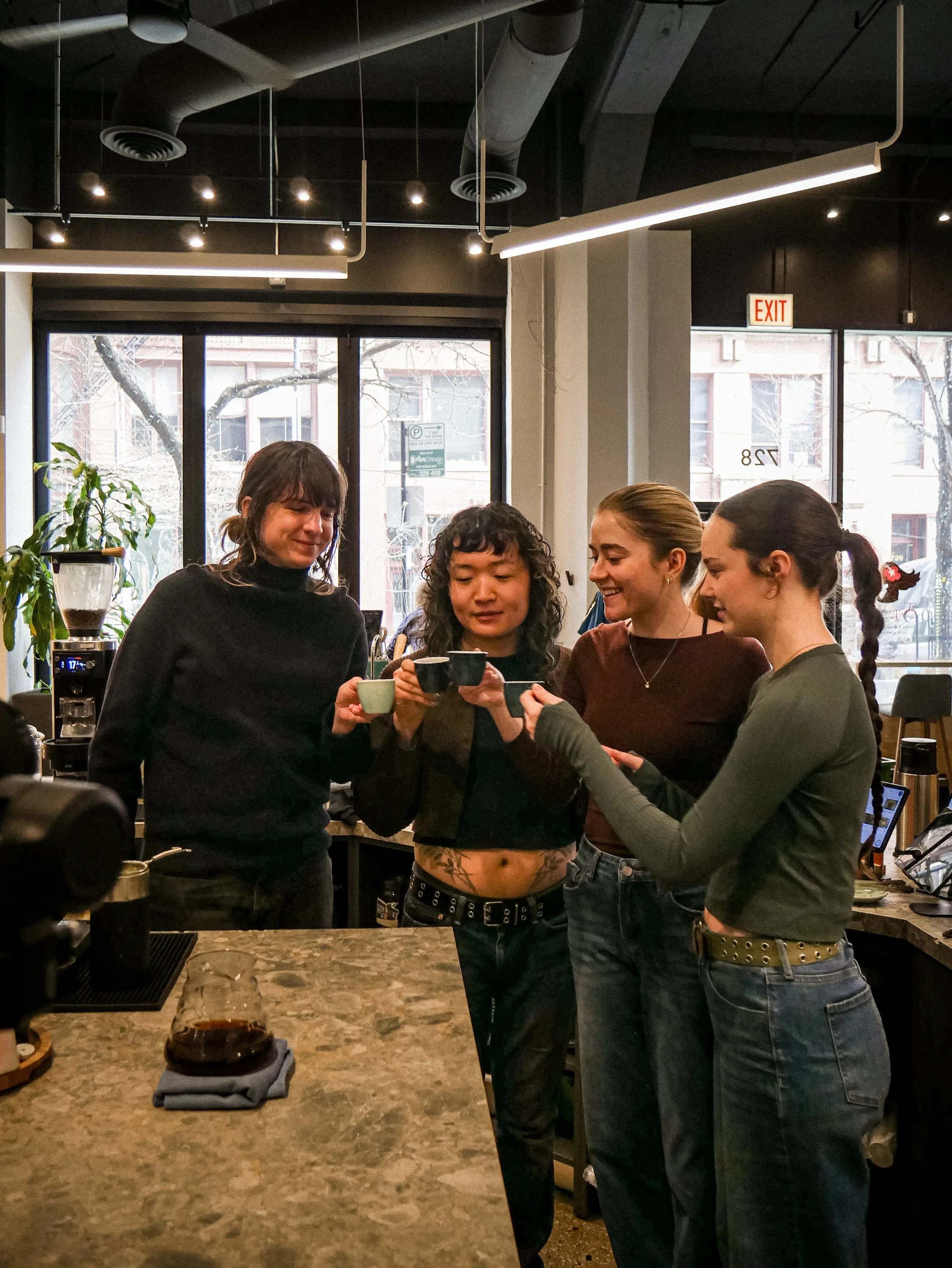Four baristas gathered around a coffee shop counter, clinking coffee cups and smiling, with one woman in the center holding a small ceramic cup and a drip coffee pot in front of her on the counter.
