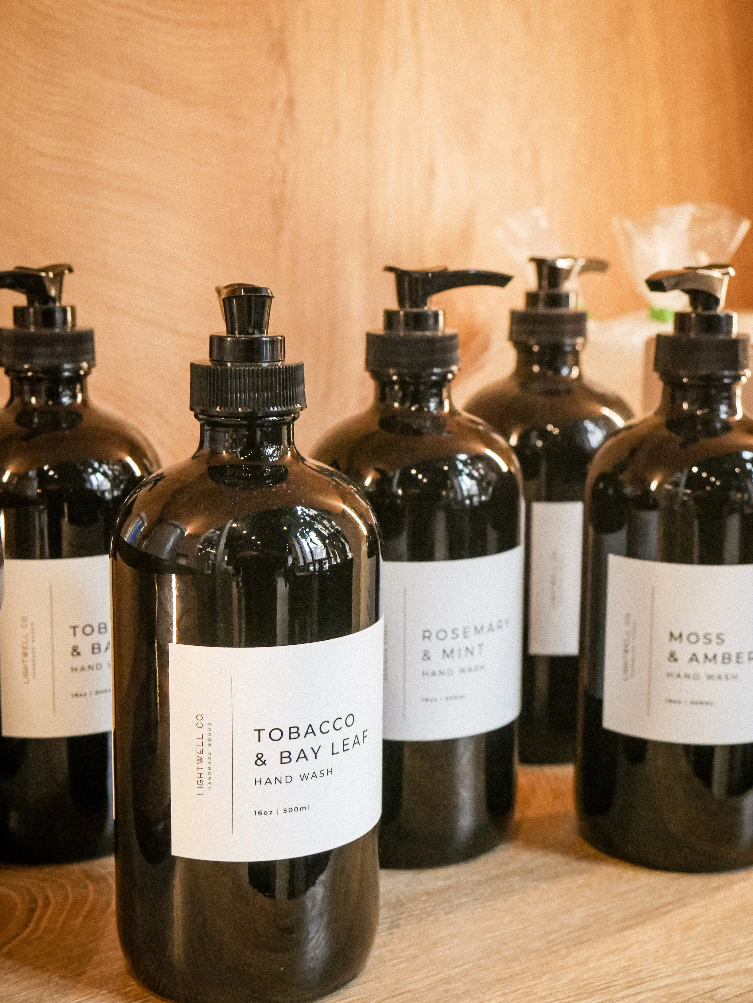 Multiple dark brown bottles of hand wash labeled with scents such as Tobacco & Bay Leaf, Rosemary & Mint, and Moss & Amber, arranged on a wooden shelf.