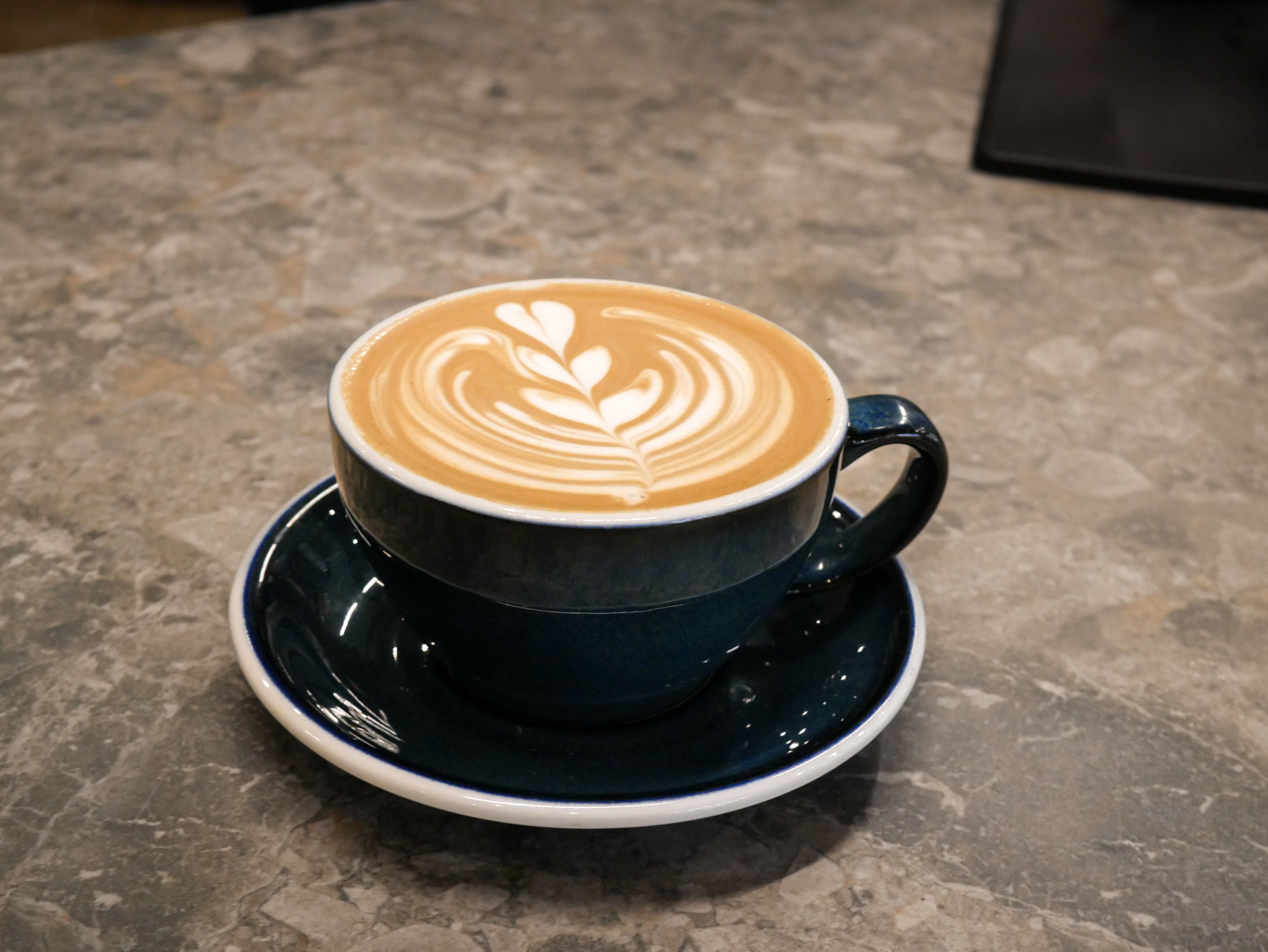 A cup of latte with heart and leaf latte art on top, placed on a matching cup and saucer on a marble table.