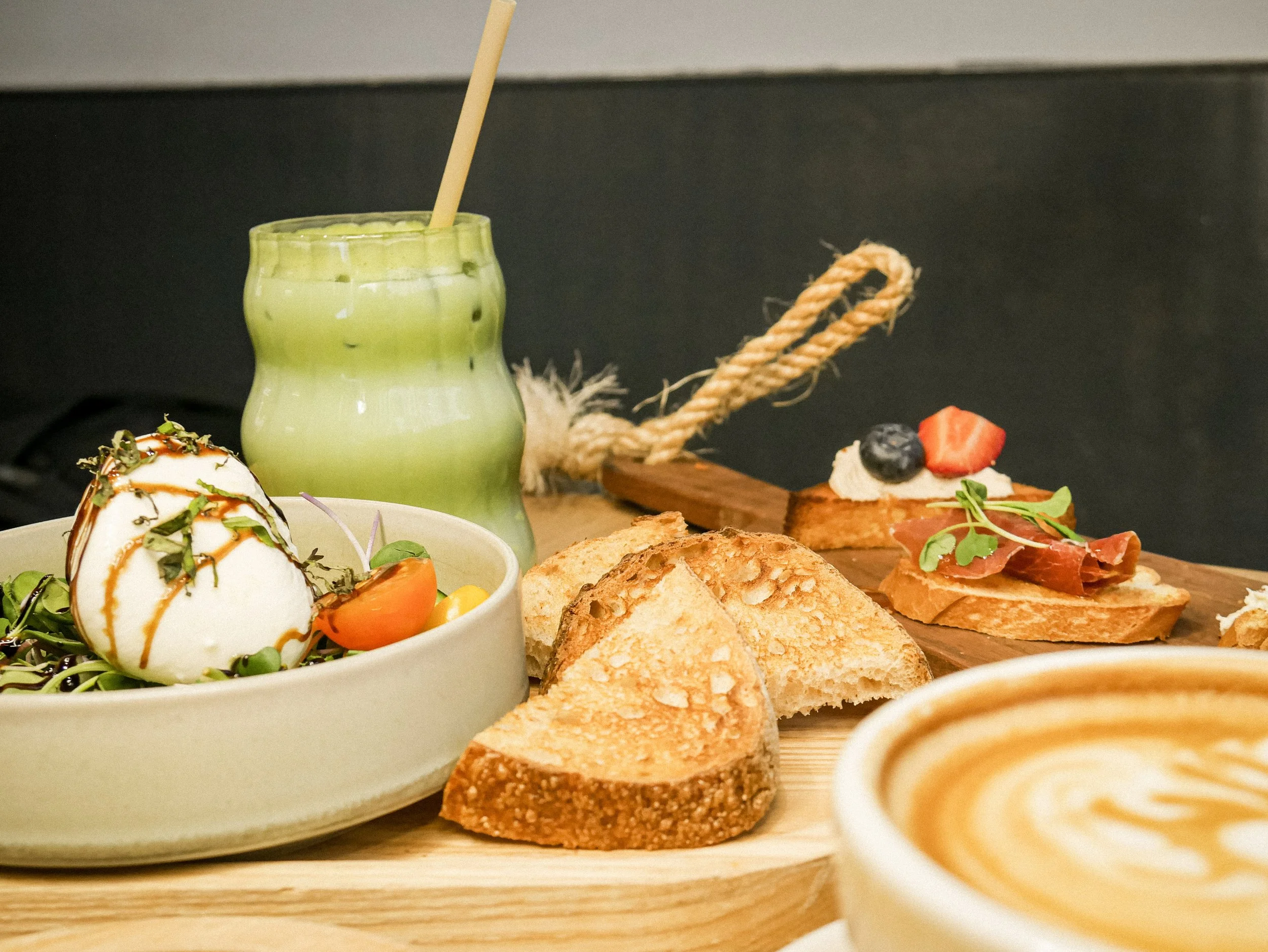 A breakfast or brunch spread with toast, prosciutto, berries, salad with mozzarella, green drink, and cappuccino with latte art.