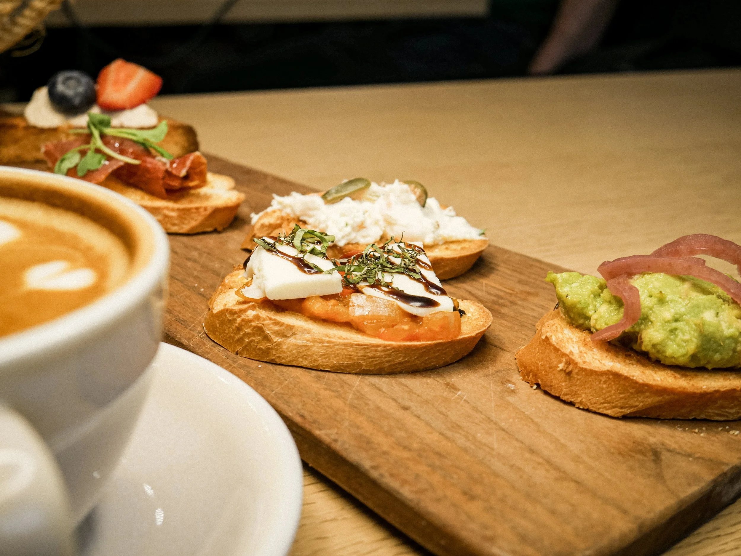 Close-up of a wooden serving board with three different types of open-faced appetizers, including a slice of bread with guacamole and pickled red onions, a slice with cheese, tomato, and herbs, and a slice with whipped cheese, meat, and crispy greens