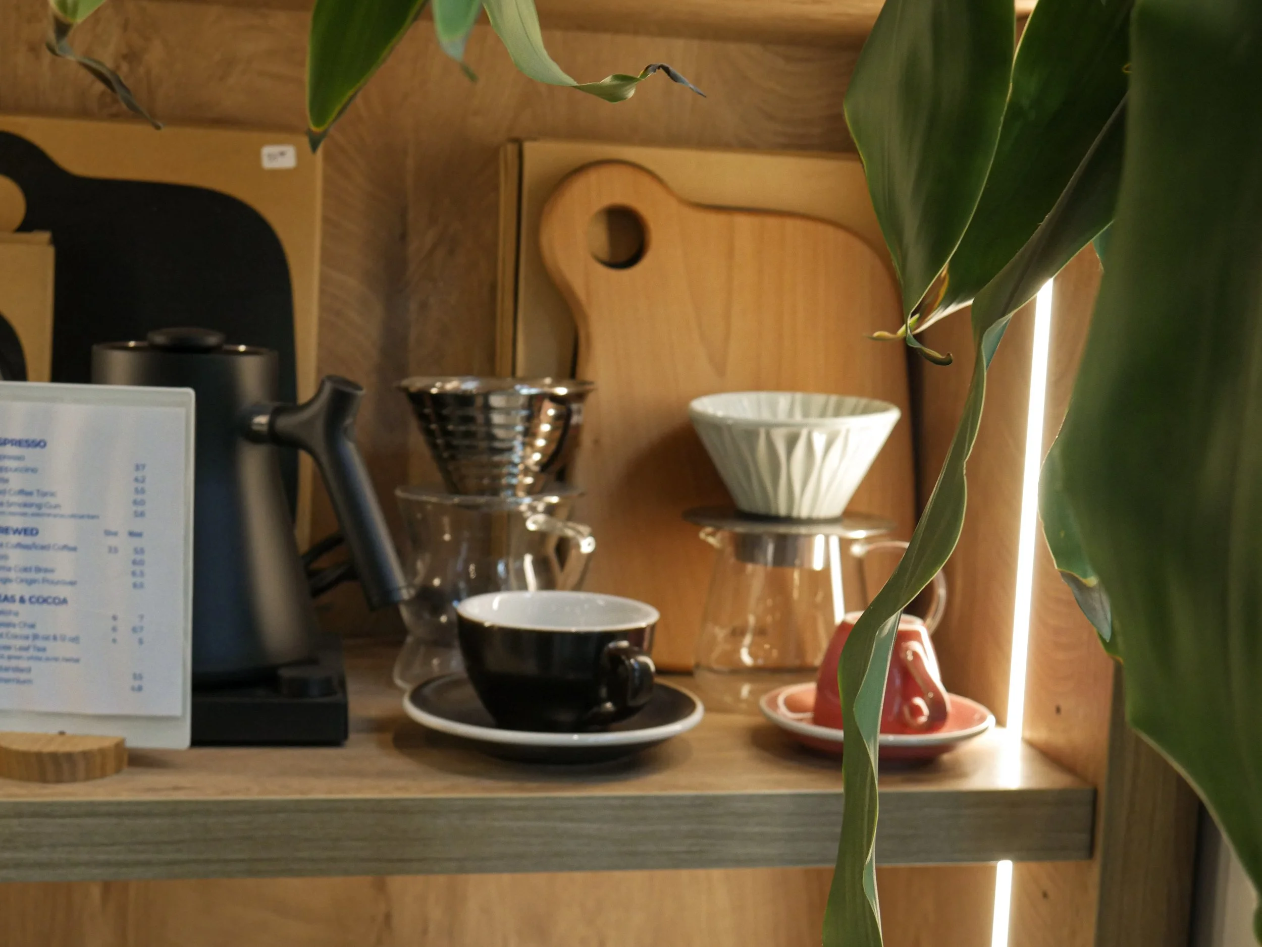 Coffee brewing setup on a wooden shelf with a black cup and saucer, a pour-over coffee dripper, glass containers, and a wooden cutting board, partially obscured by large green leaves.