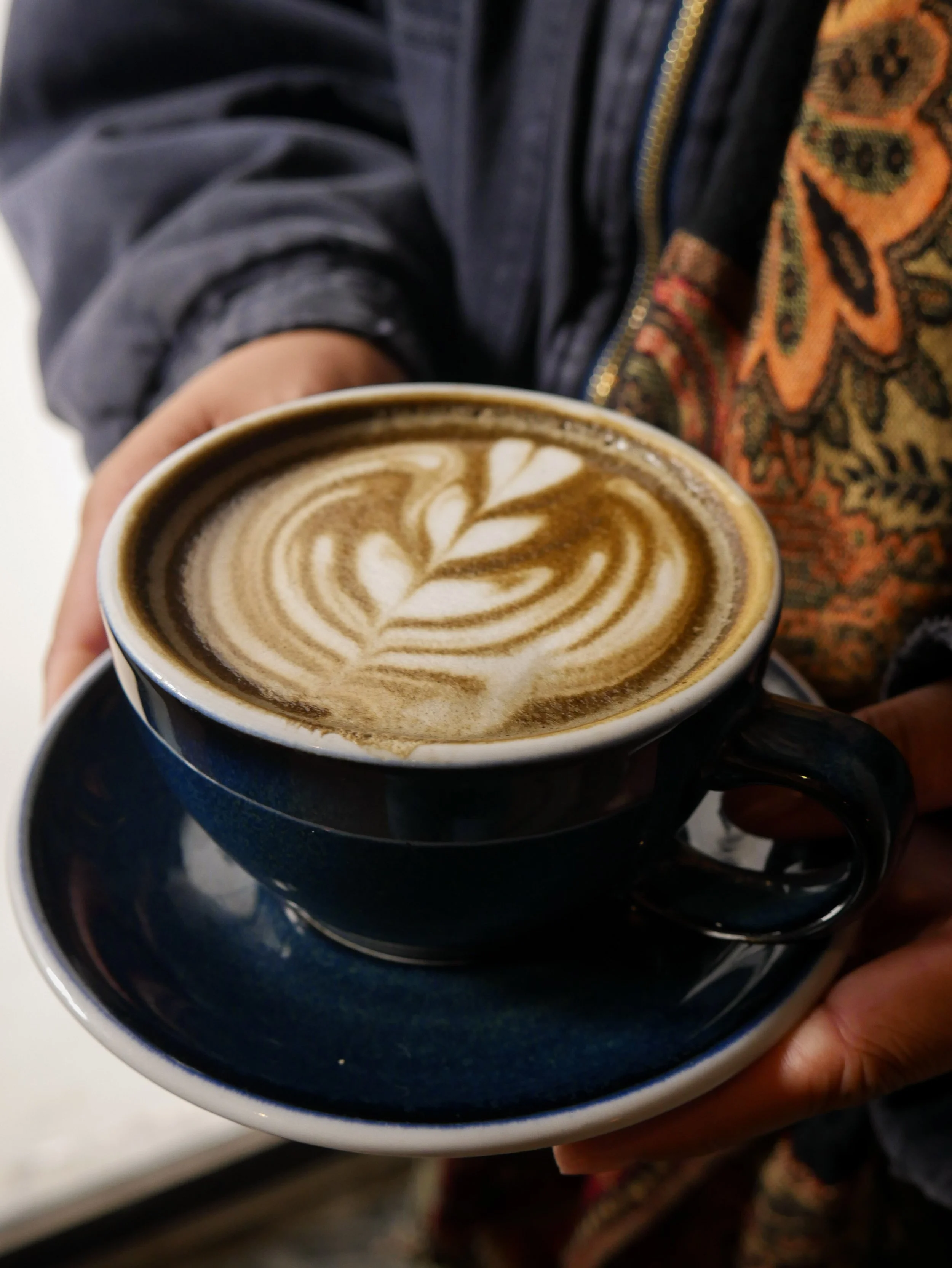 Person holding a dark blue cup of coffee with latte art in a matching saucer, with a floral or patterned scarf and a jacket partially visible.