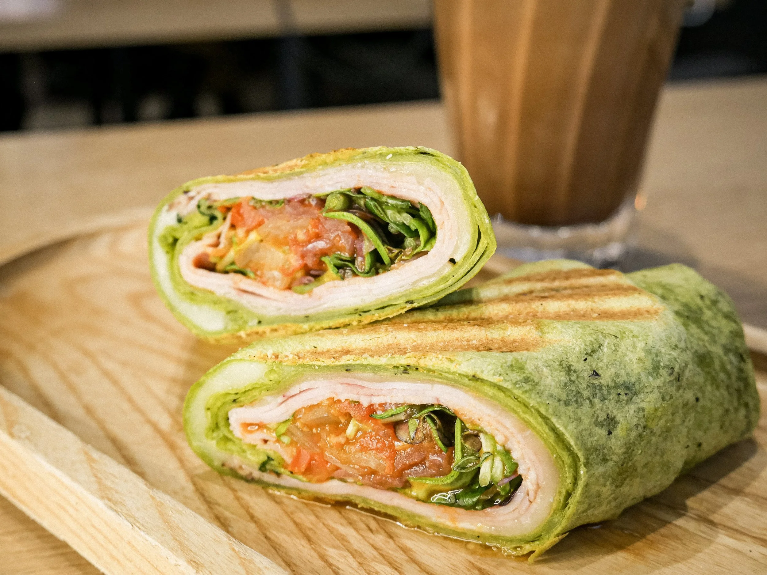 A sliced vegetable wrap with grilled green tortilla, showing layers of sliced turkey, vegetables, and a romaine lettuce base, served on a wooden board with a drink in the background.