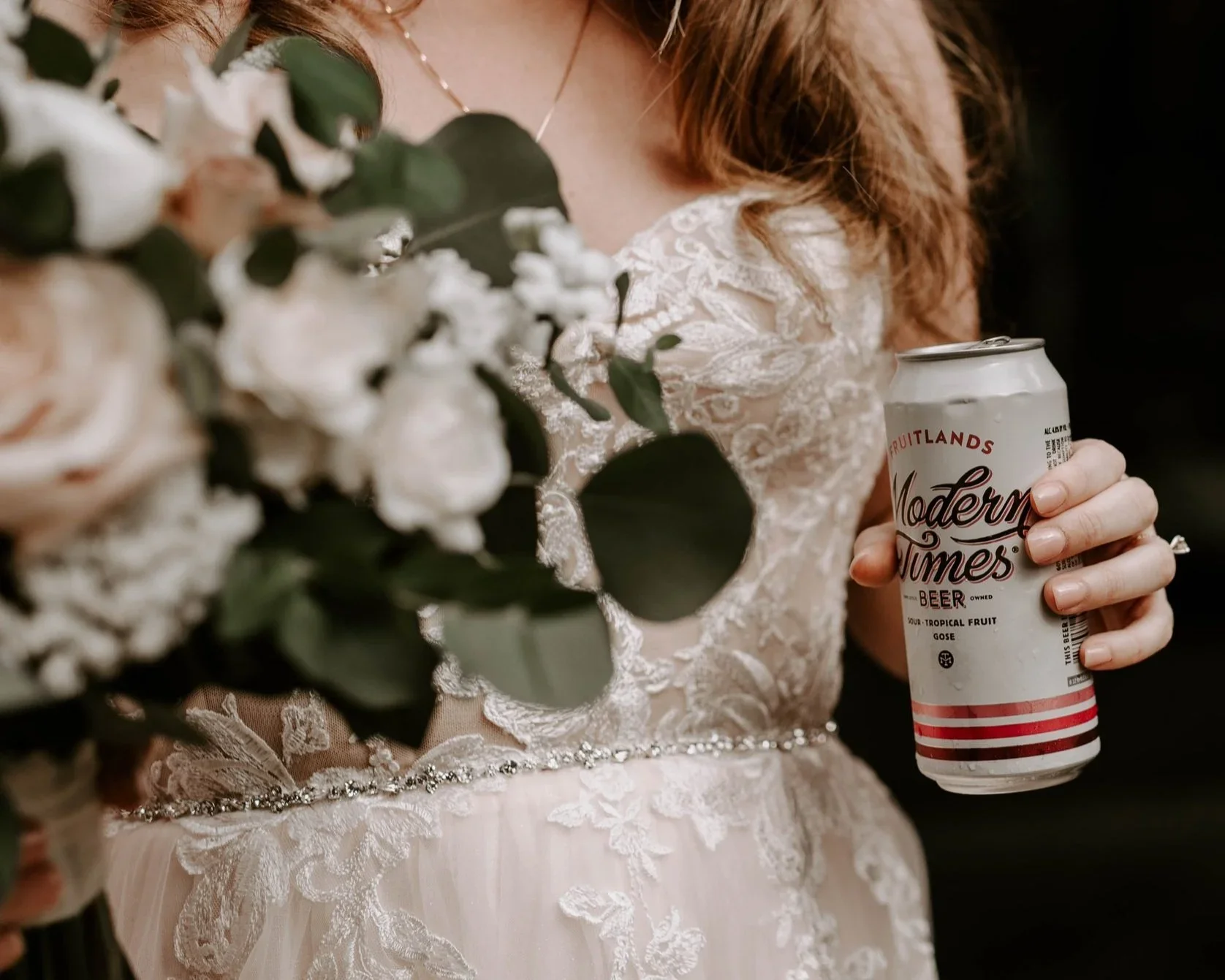 A woman in a detailed white lace wedding dress holding a bouquet of white flowers with greenery and a can of Modern Times beer.