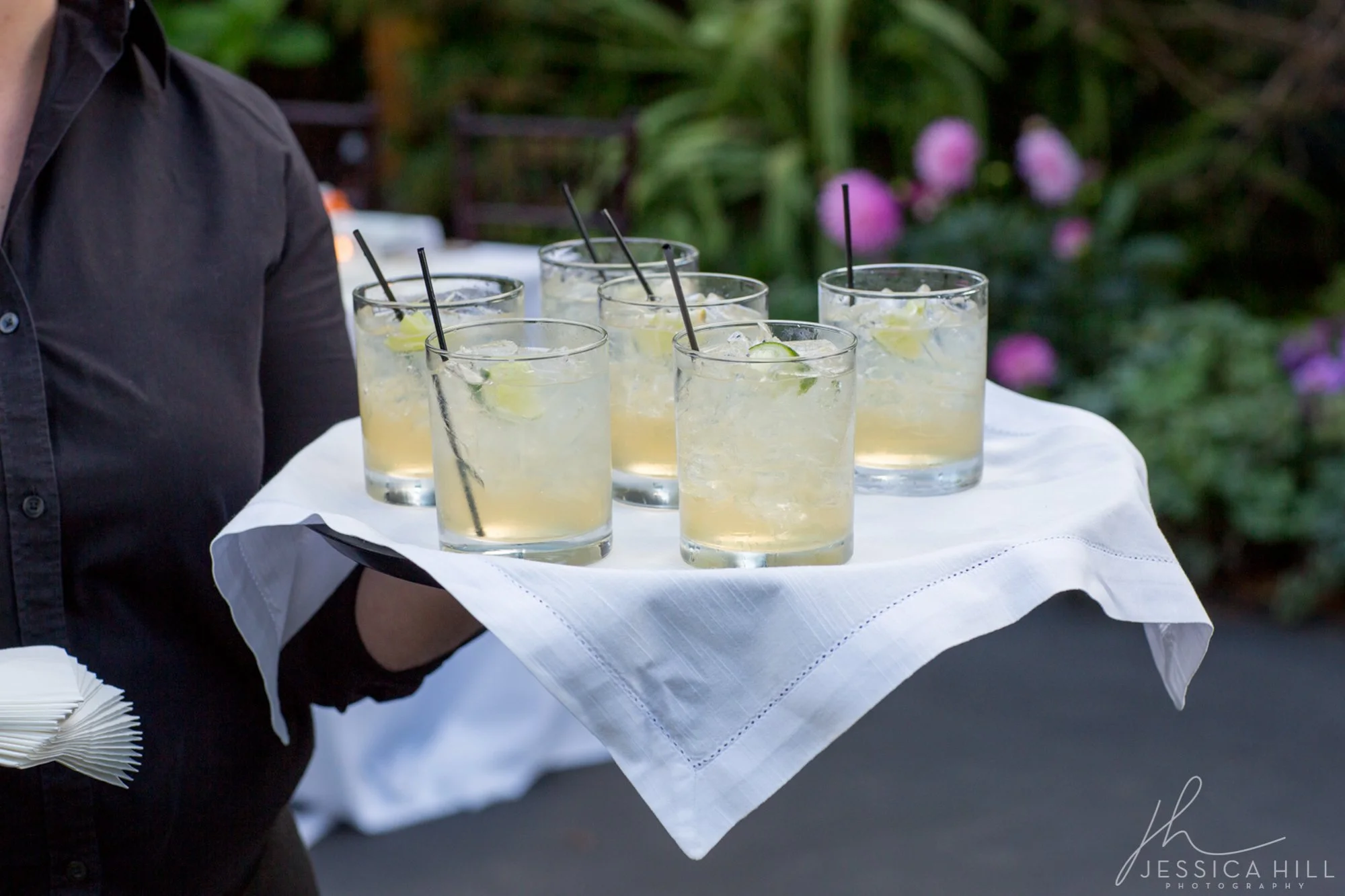 A person holding a tray with six glasses of lemonade garnished with lime slices and black straws, set on a white cloth.