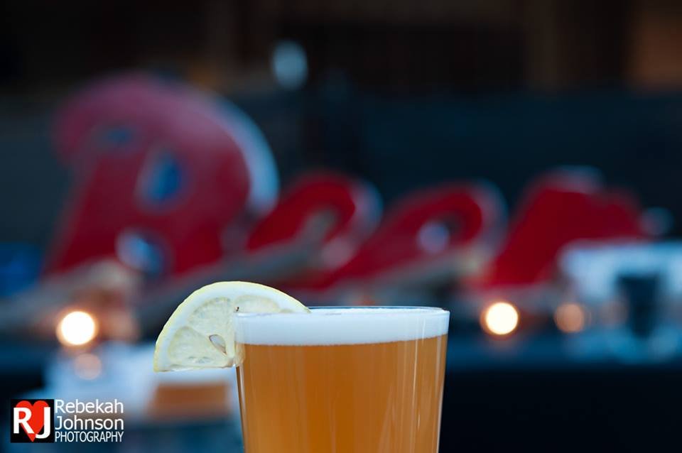 A glass of beer with a lemon wedge garnish in the foreground, blurred background with red and black decorations and soft lights.