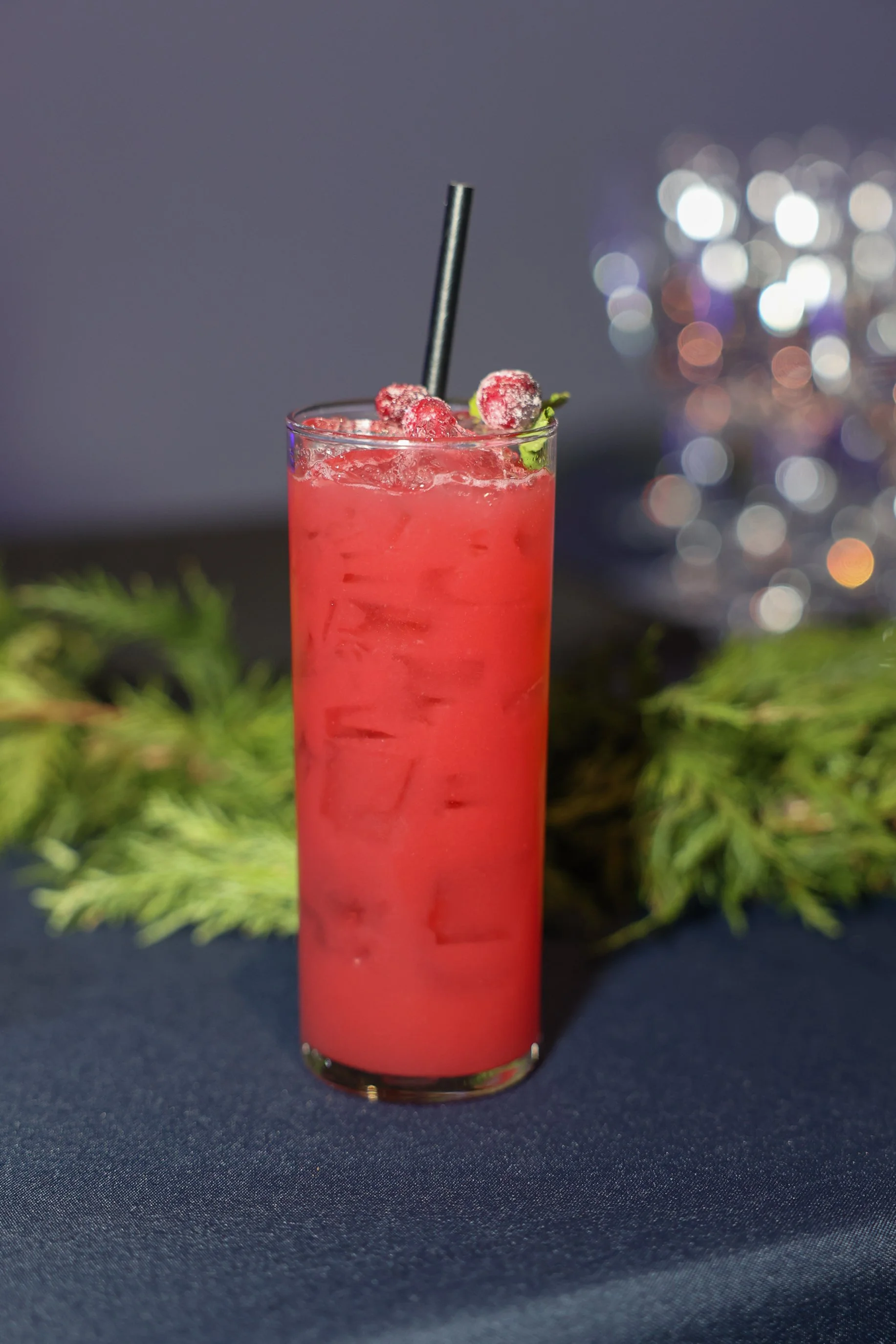A tall glass of pink cranberry cocktail garnished with frozen cranberries and a mint sprig, with a black straw, on a dark surface with a blue background and blurred sparkling lights in the background.