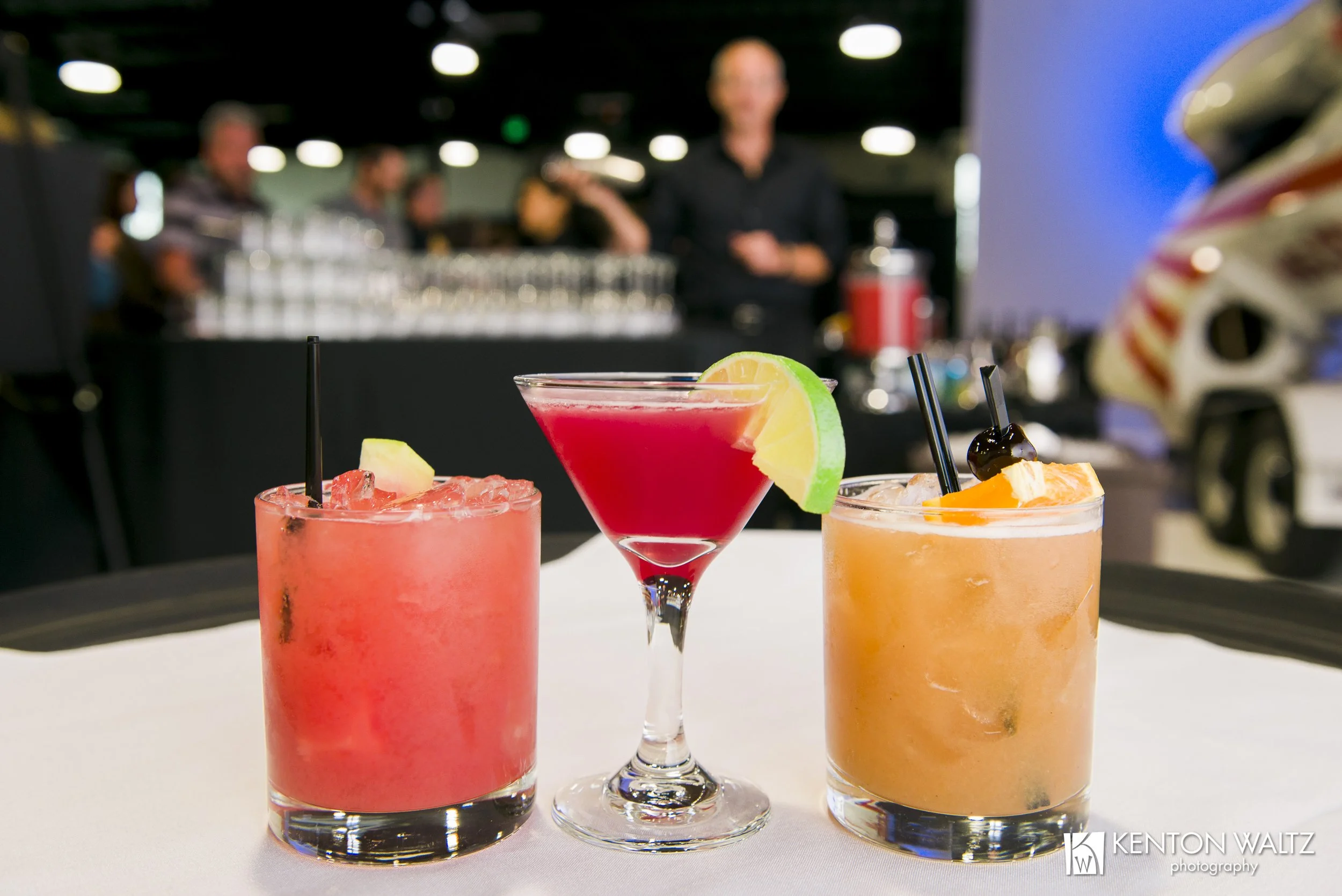 Three colorful cocktails with garnishes on a white table at a bar or restaurant, with people in the background.