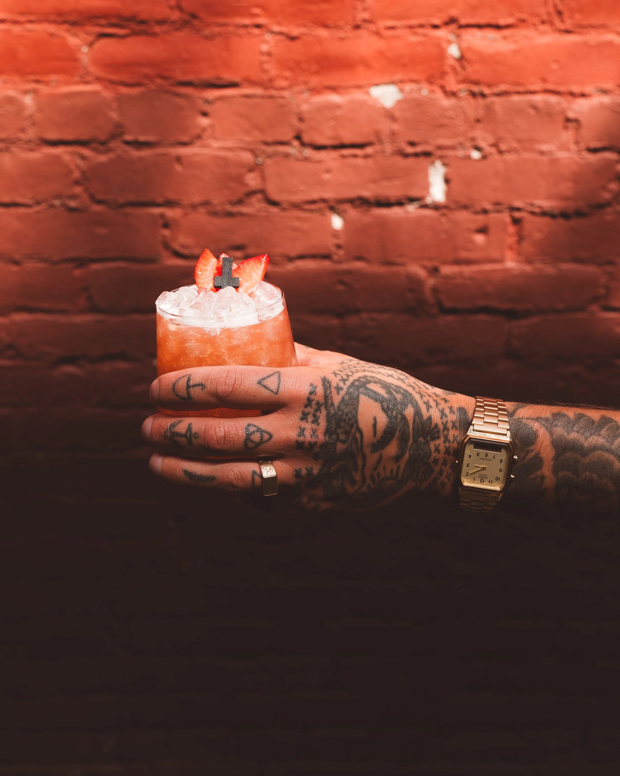 A person with tattoos on their hand and wrist is holding a strawberry cocktail drink topped with sliced strawberries and a small black cross umbrella, against a red brick wall background.