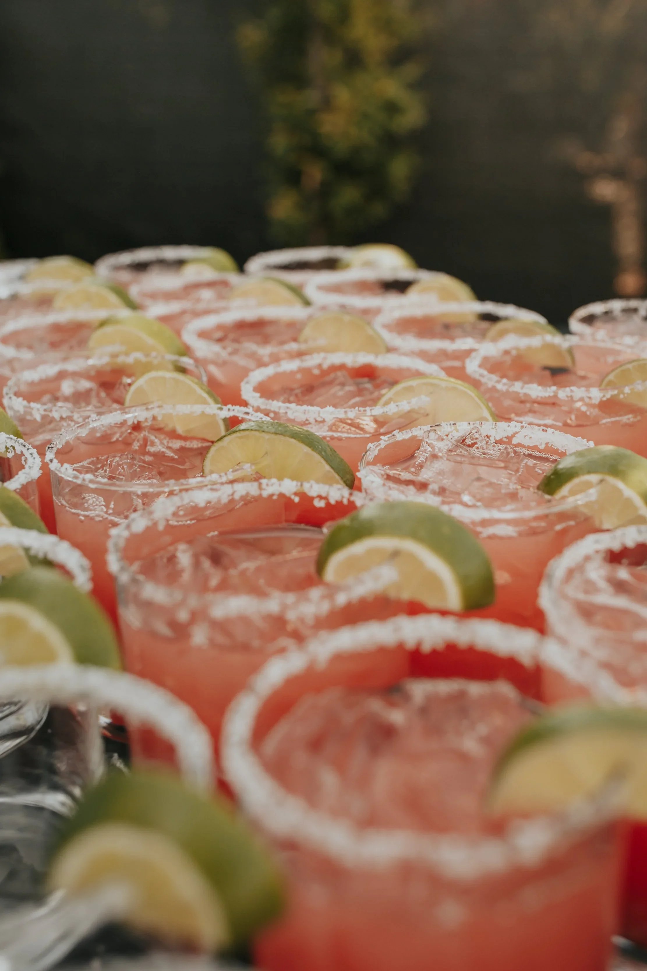 A close-up of pink cocktails in glasses garnished with lime wedges and salted rims, arranged outdoors with blurred trees in the background.