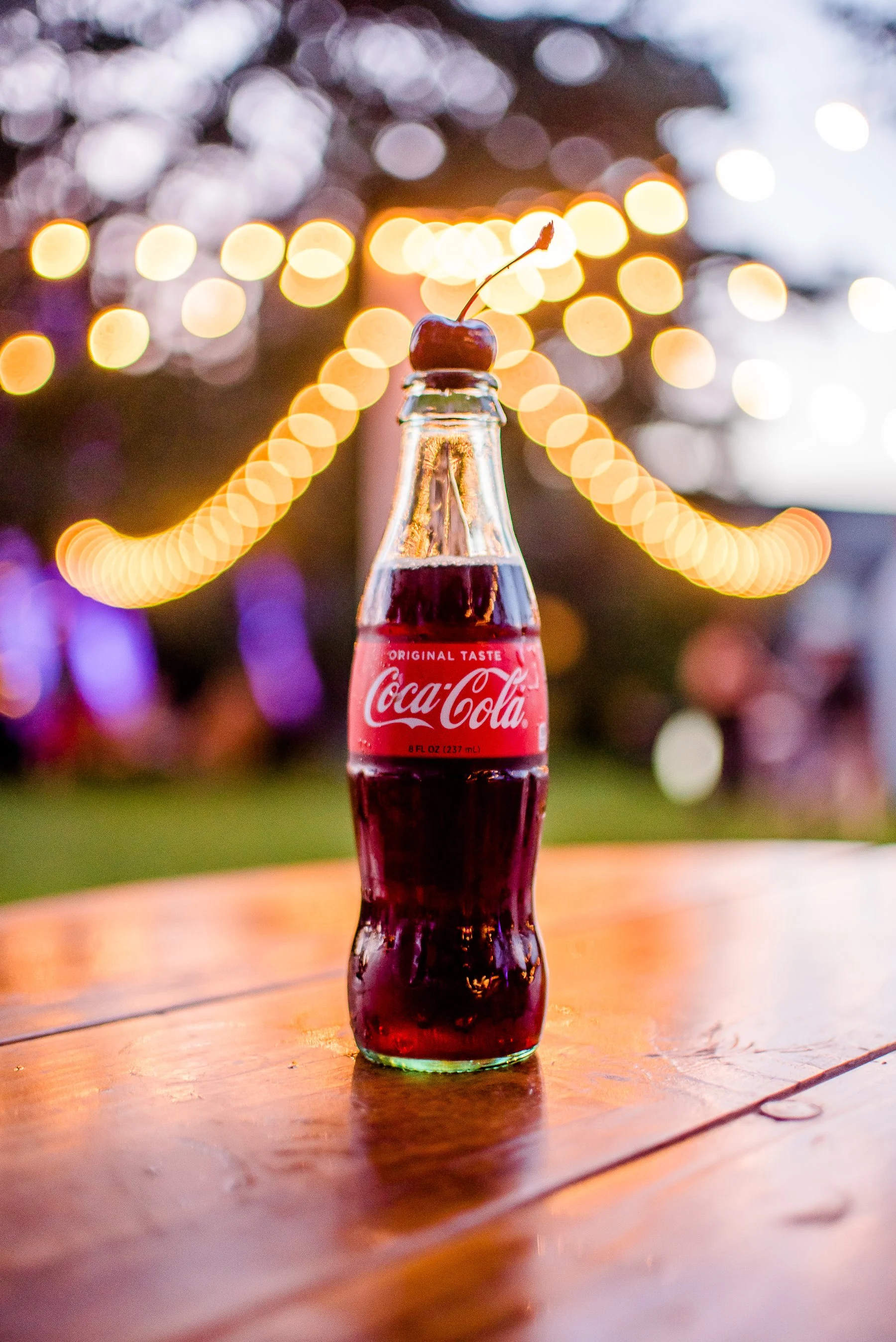 A glass bottle of Coca-Cola with a cherry on top, placed on a wooden table outdoors during evening with blurred lights and trees in the background.