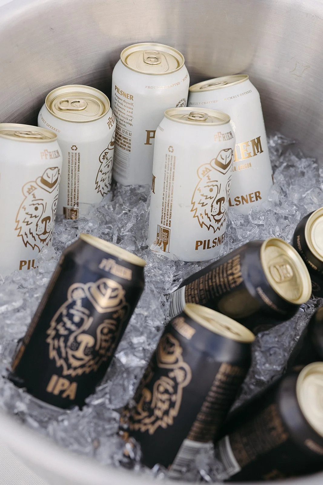 Cans of beer in a cooler filled with ice. The cans are mainly white with some black, featuring a lion logo and the word 'Pilsner' and 'IPA'.