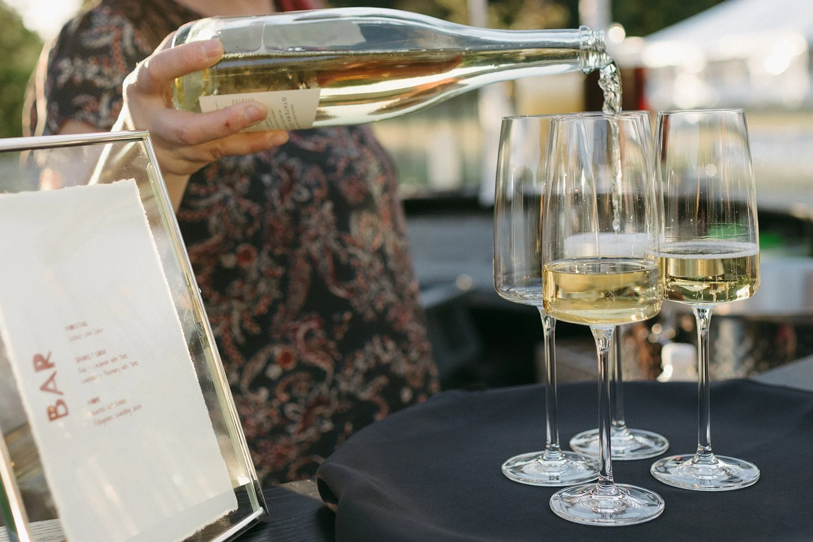 Person pouring white wine into wine glasses at an outdoor event, with a menu or sign in the foreground.