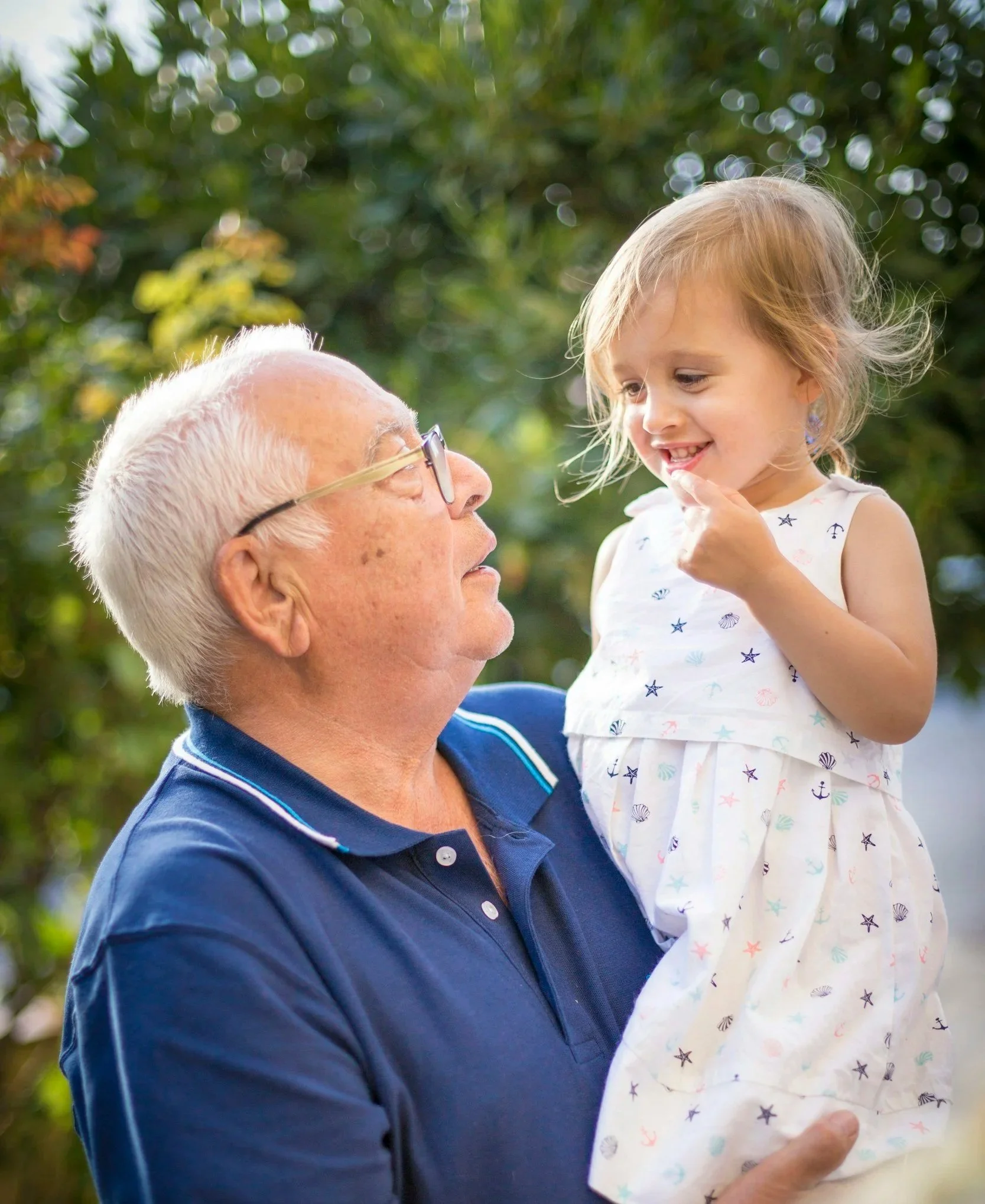 An elderly man with glasses holding a young girl, dressed in a white dress with nautical patterns, in a park with lush greenery in the background.