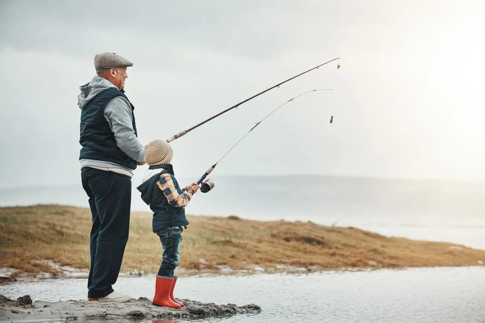 A grandpa and a young boy fishing by a body of water on a cloudy day