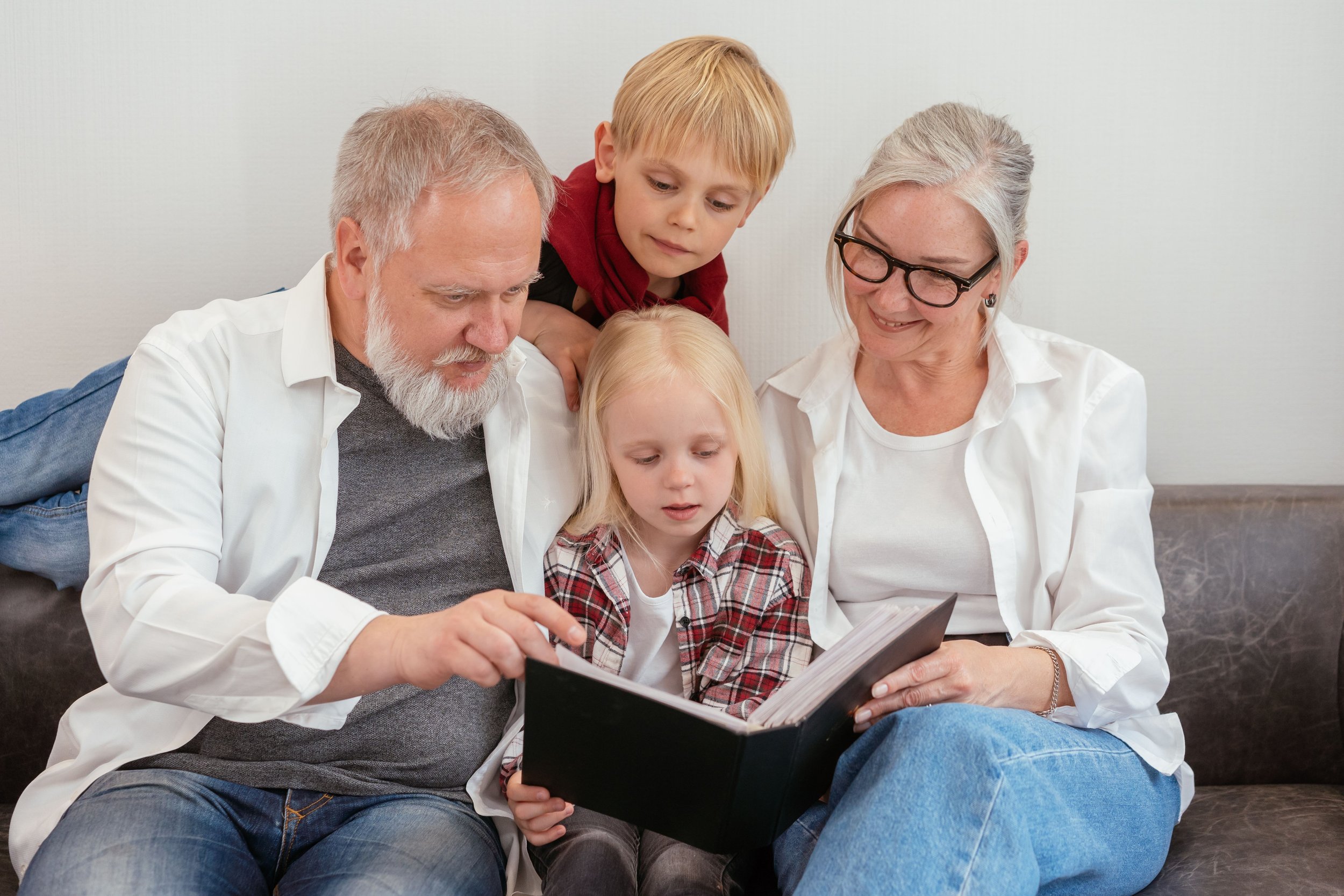 A multigenerational family, including an elderly man and woman, a middle-aged man and woman, and two young children, gathered around looking at a photo album together in a cozy room.