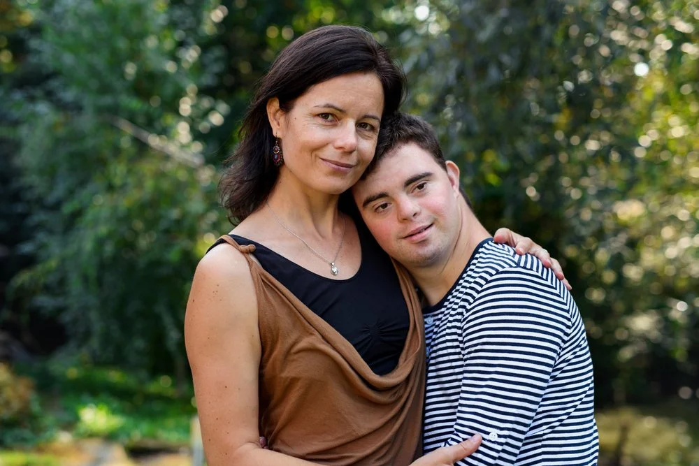 A woman and a young man with special needs hugging outdoors with greenery in the background.