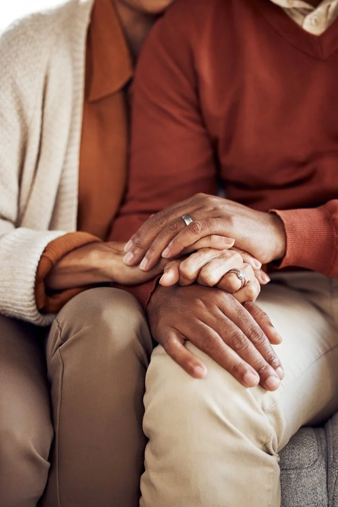 Close-up of two elderly black people holding hands, two with wedding rings, sitting closely together.
