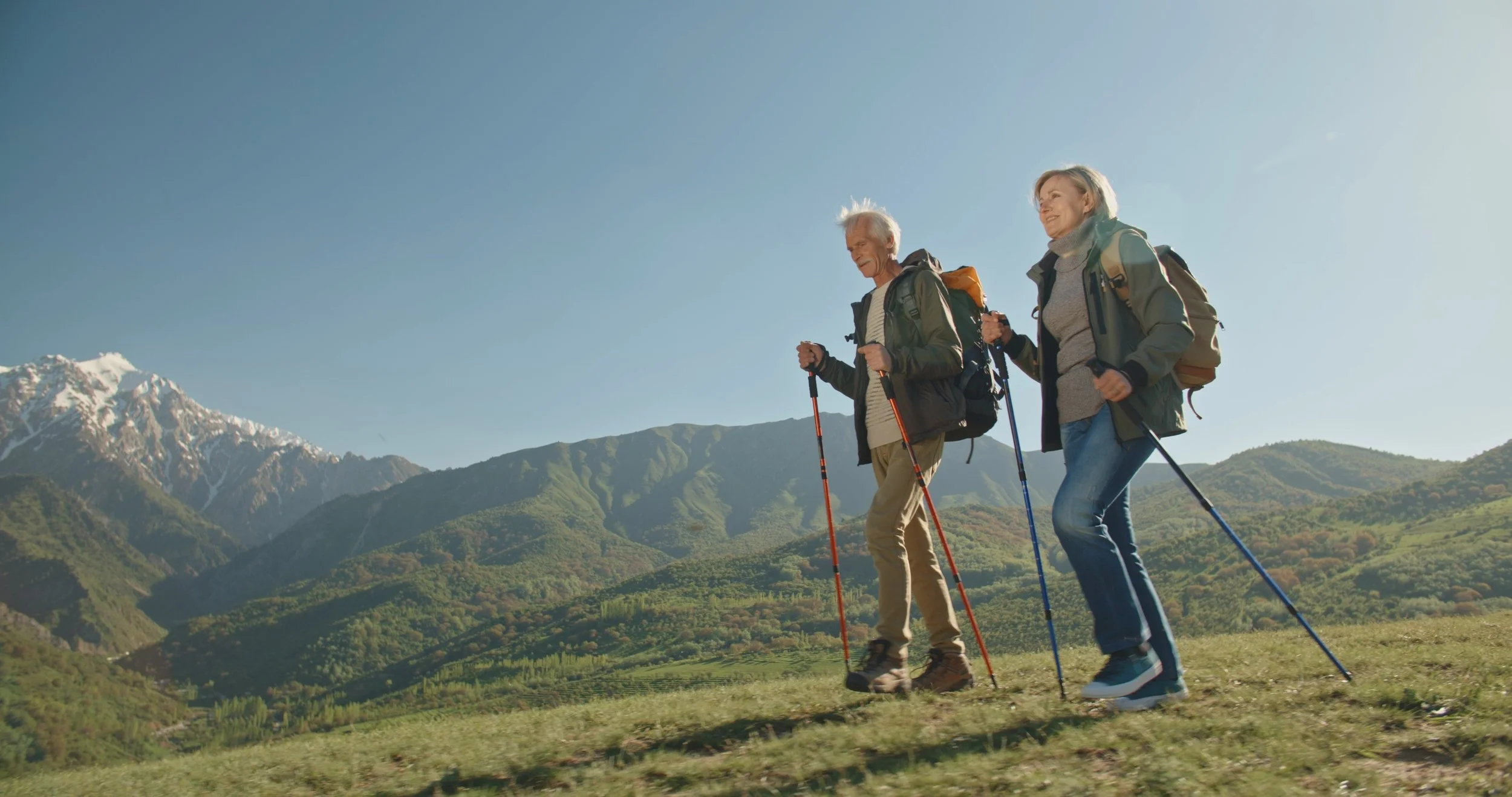 A retired couple hiking with trekking poles in a scenic mountainous landscape with snow-capped peaks and green hills under a clear blue sky.