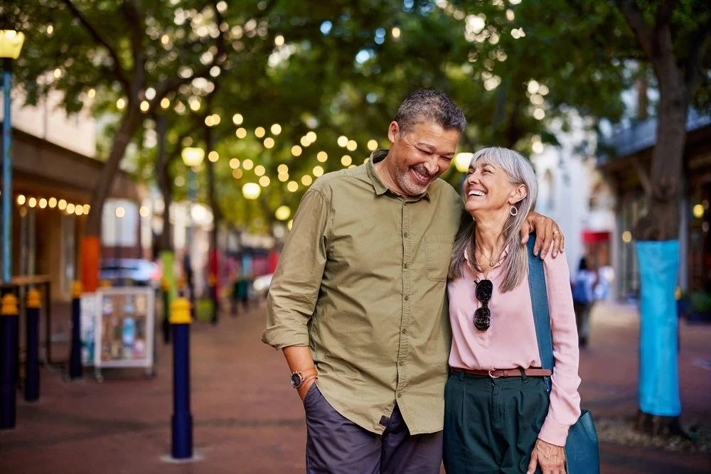 A middle-aged man and woman sharing a laugh and walking arm in arm on a city street with trees and string lights, during early evening.