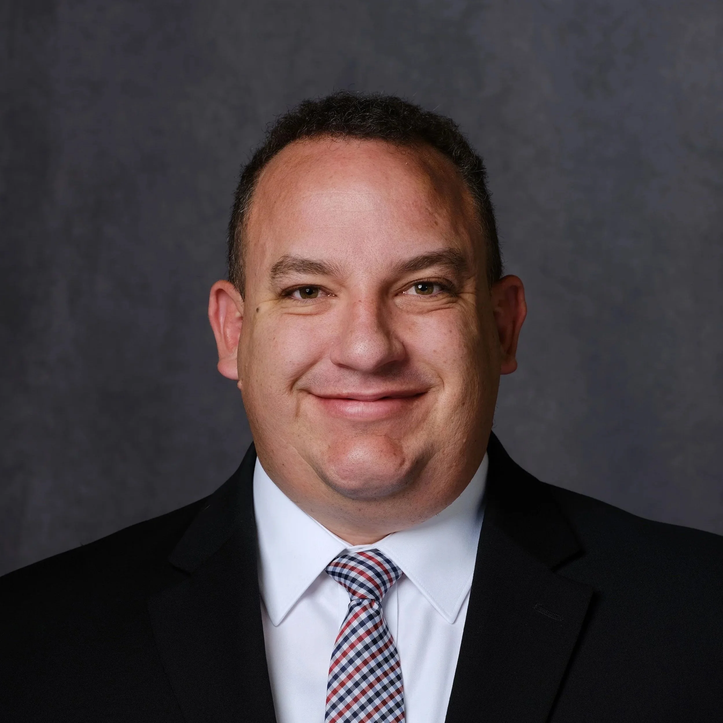 Close-up of a man's face, showing his eyes, forehead, and part of his nose and mouth, dressed in a black suit, white shirt, and a checkered tie, with a dark gray background.