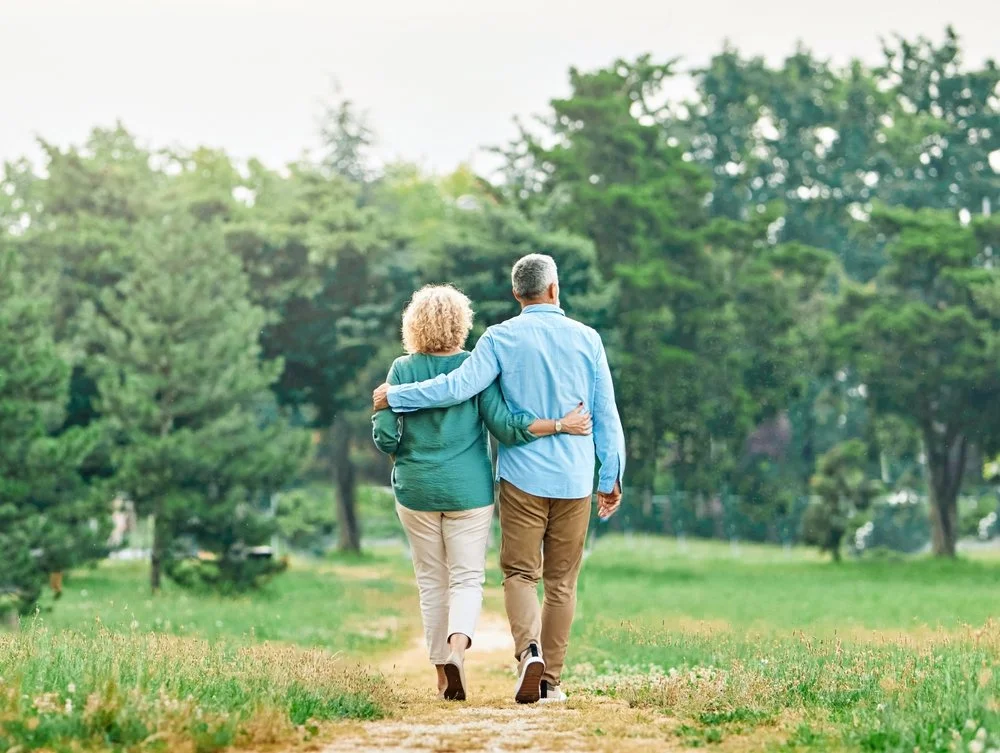 A retired couple walking together arm in arm in a park, surrounded by green trees and grassy area.