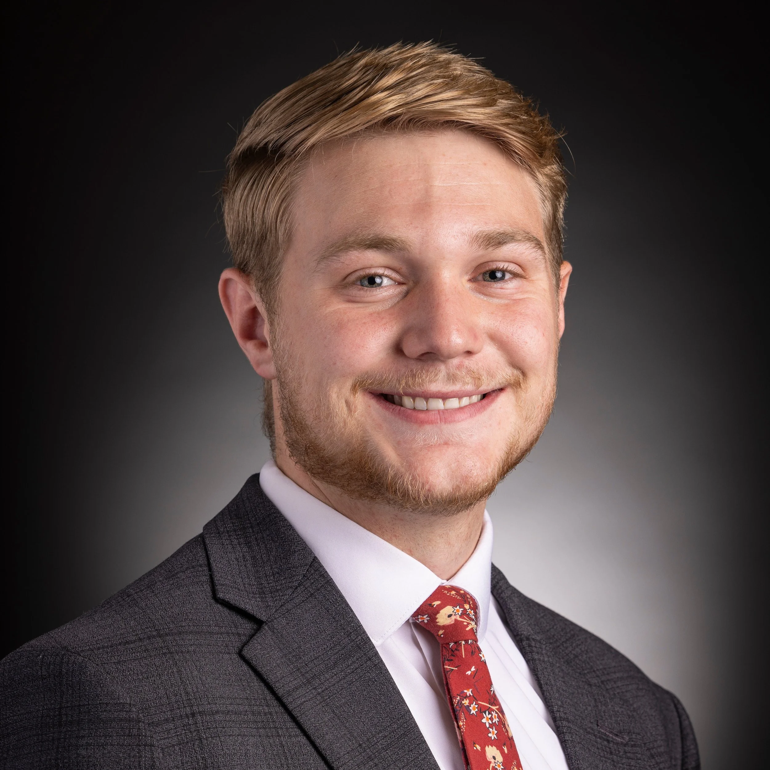 Close-up portrait of a young financial advisor with light brown hair, blue eyes, wearing a dark gray patterned suit, white shirt, and a red tie with floral design, smiling against a dark background.