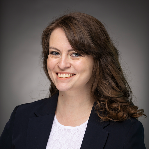 A professional woman with long brown hair and a friendly smile, wearing a dark blazer and a white top, posing against a plain gray background.