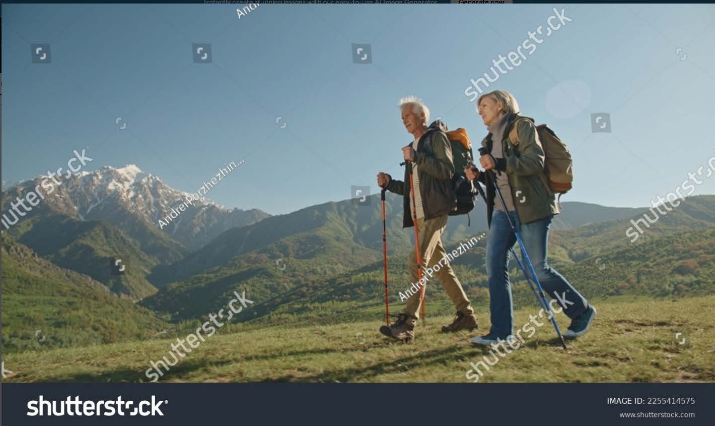 Two seniors hiking on a mountain trail with snow-capped peaks in the background on a clear day.