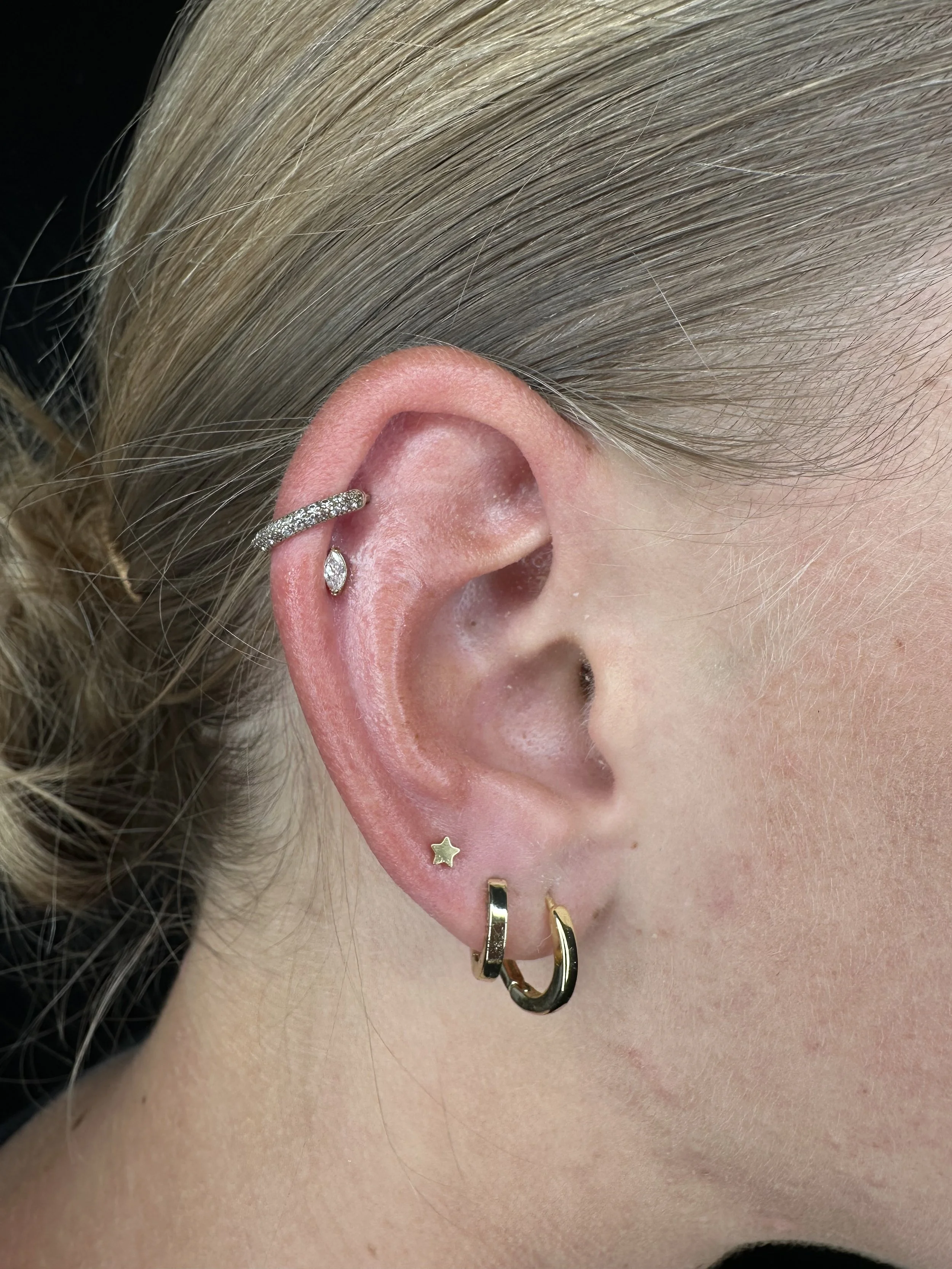 Close-up of a person's ear with multiple earrings, including a diamond-studded hoop, a small star-shaped earring, a small diamond-shaped earring, a gold hoop, and a black and gold curved earring.