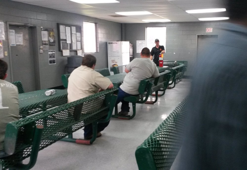 Three men seated at green benches in a room with grey walls, listening to a woman instructor demonstrating fire safety with a fire extinguisher.