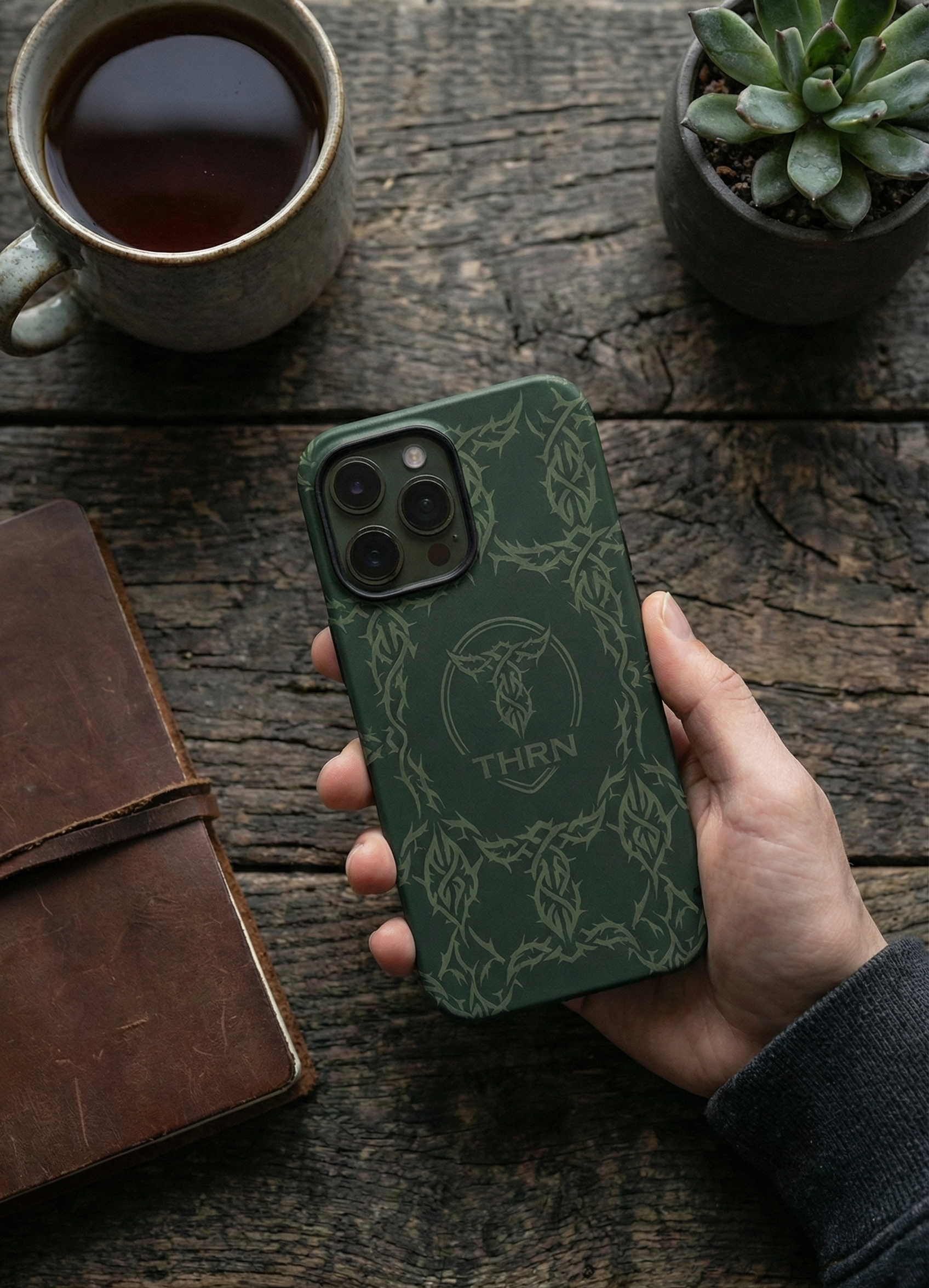 A hand holding a green phone with a tribal logo and the word 'THRN' on a rustic wooden table. Surrounding items include a cup of coffee, a small potted succulent plant, and a closed brown leather notebook.