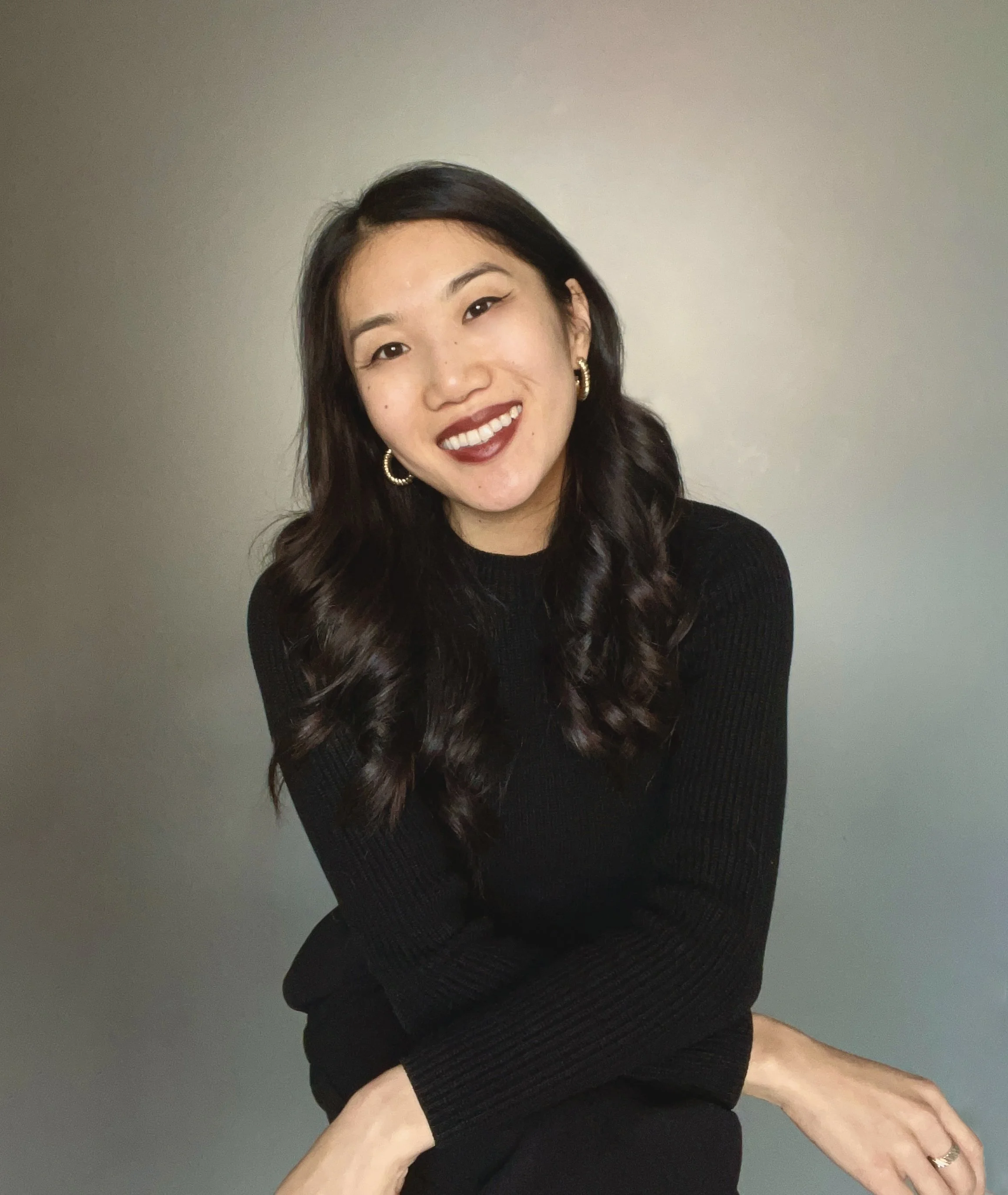 A smiling woman with dark hair, wearing gold hoop earrings and a black long-sleeve top, seated against a plain light gray background.