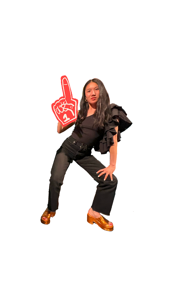 Young woman posing with a large red foam finger that has a white hand making a peace sign, wearing black ruffled top, black pants, and gold platform shoes, against a black background.