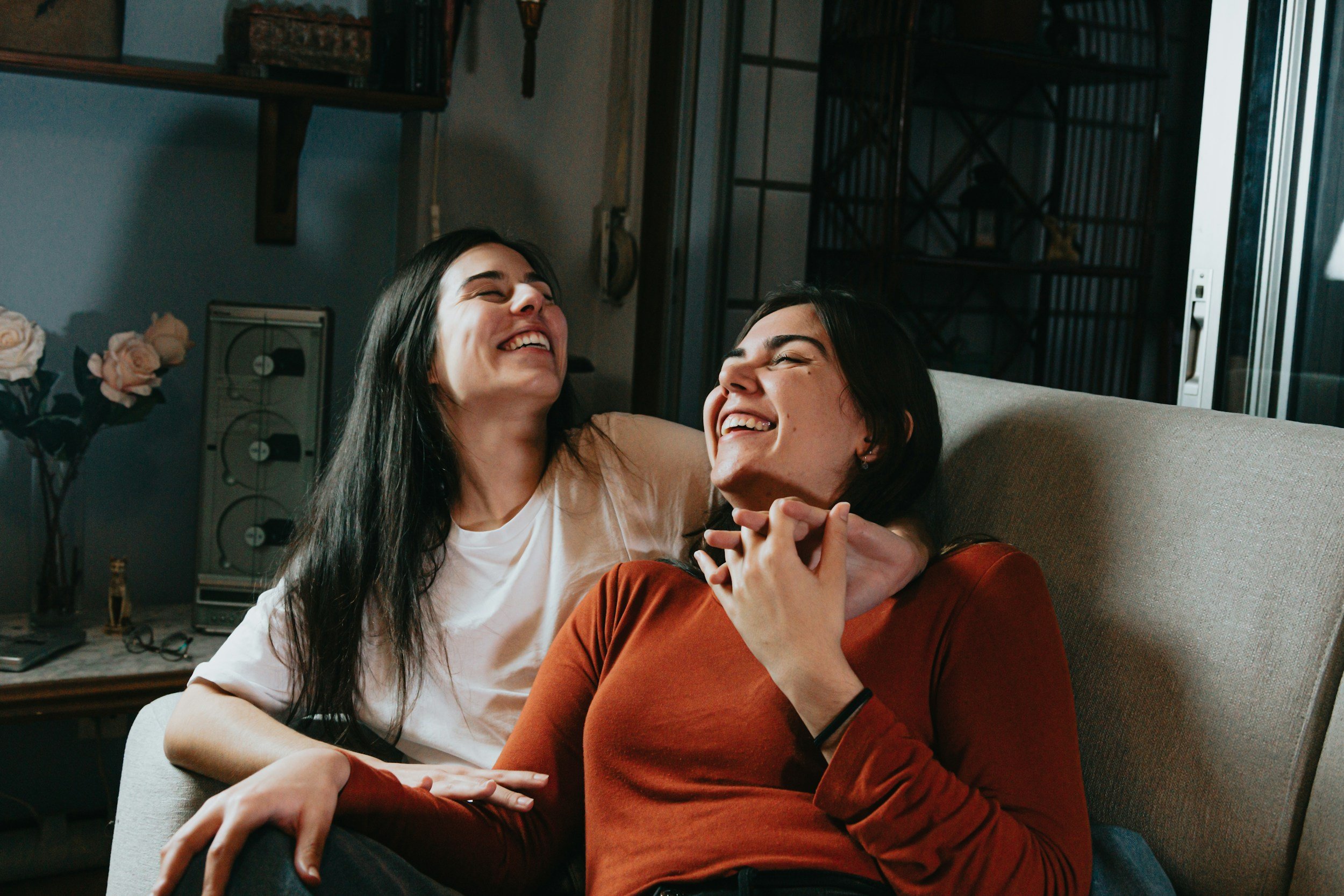 Two women sitting on a sofa, laughing and smiling, one with long black hair and white shirt, the other with short dark hair and brown top, in a cozy living room.
