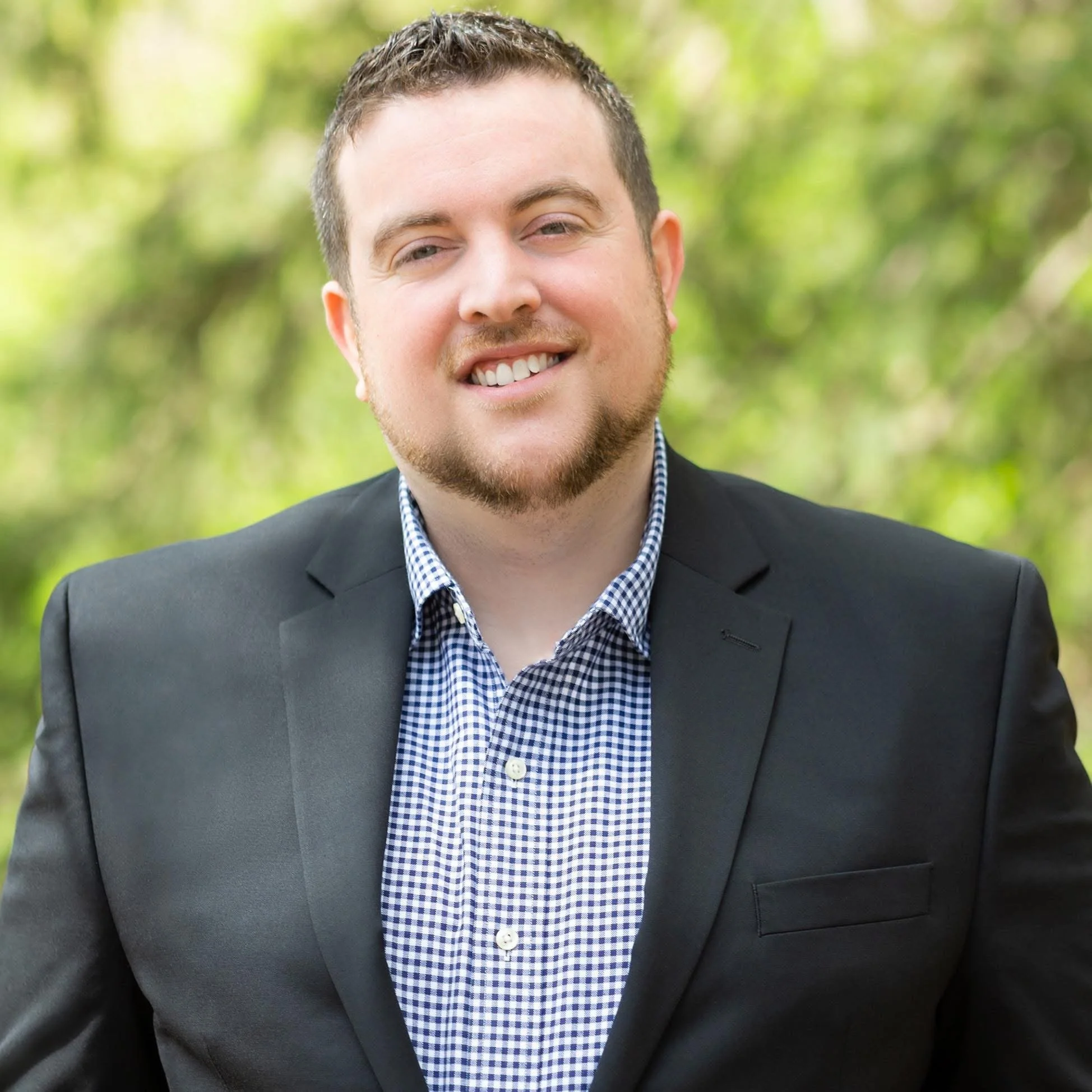 A young man with short brown hair and a beard, wearing a black suit jacket and a blue checkered dress shirt, smiling outdoors with a blurred green background.