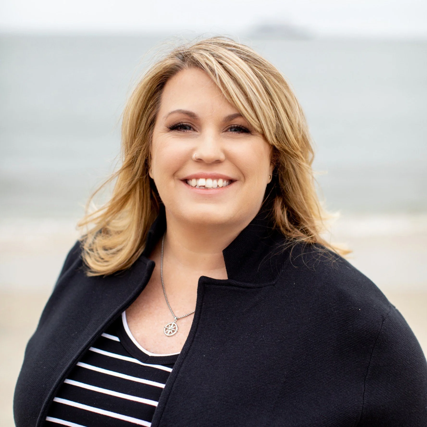 A woman with shoulder-length blonde hair smiling at the camera on a beach, wearing a black jacket and a striped shirt.