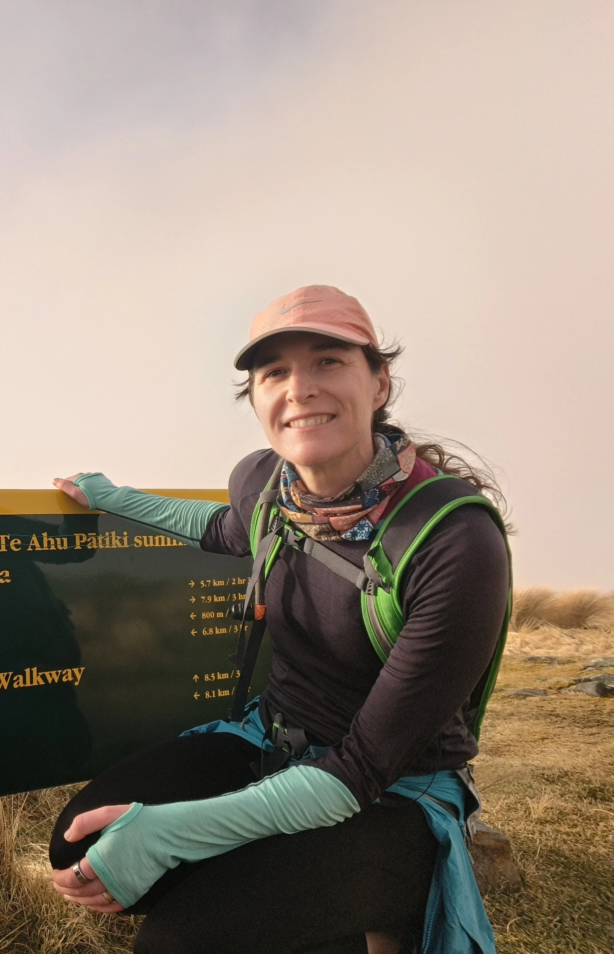 A woman outdoors, smiling, wearing a pink cap, colorful neck gaiter, black jacket, teal and black leggings, and a green backpack. She is holding a black sign with yellow text, and there is a natural landscape with grass and cloudy sky in the background.